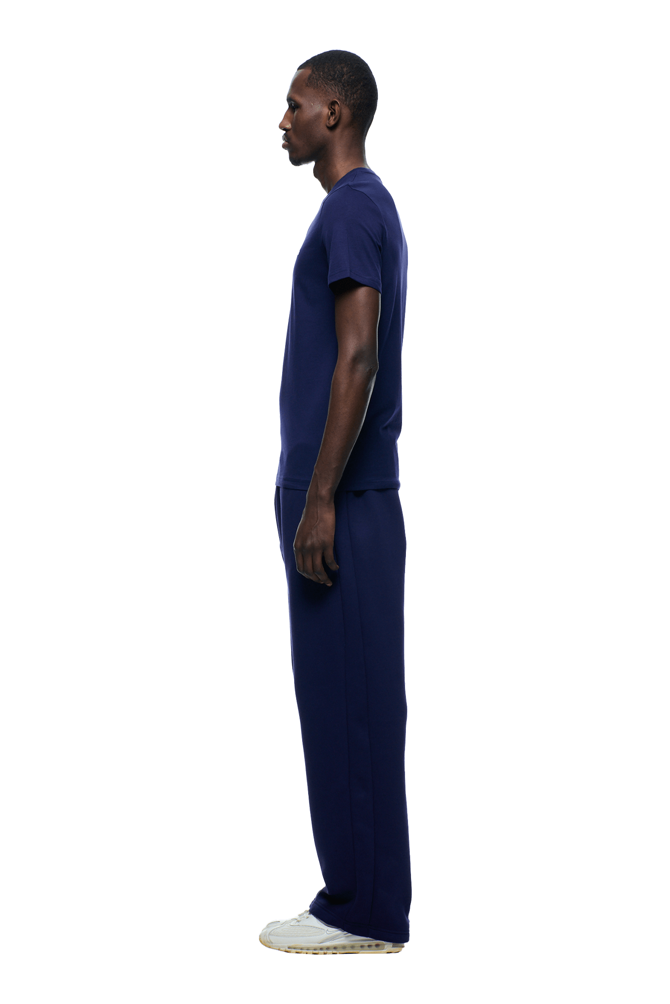 A man stands in profile facing left, wearing the 6PM SLIM FIT T-SHIRT ORBIT BLUE with matching pants and white sneakers. Made from premium cotton for everyday structure. The background is transparent.