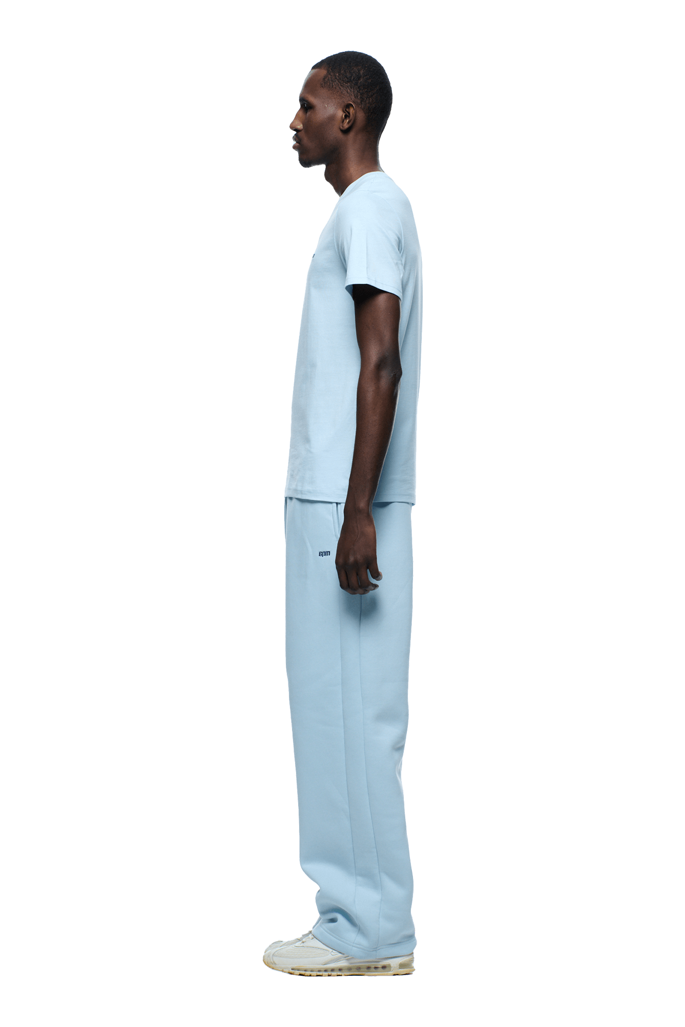 A man stands in profile, facing left, wearing the 6PM SLIM FIT T-SHIRT ICE BLUE, paired with loose sweatpants and white sneakers, set against a plain background.