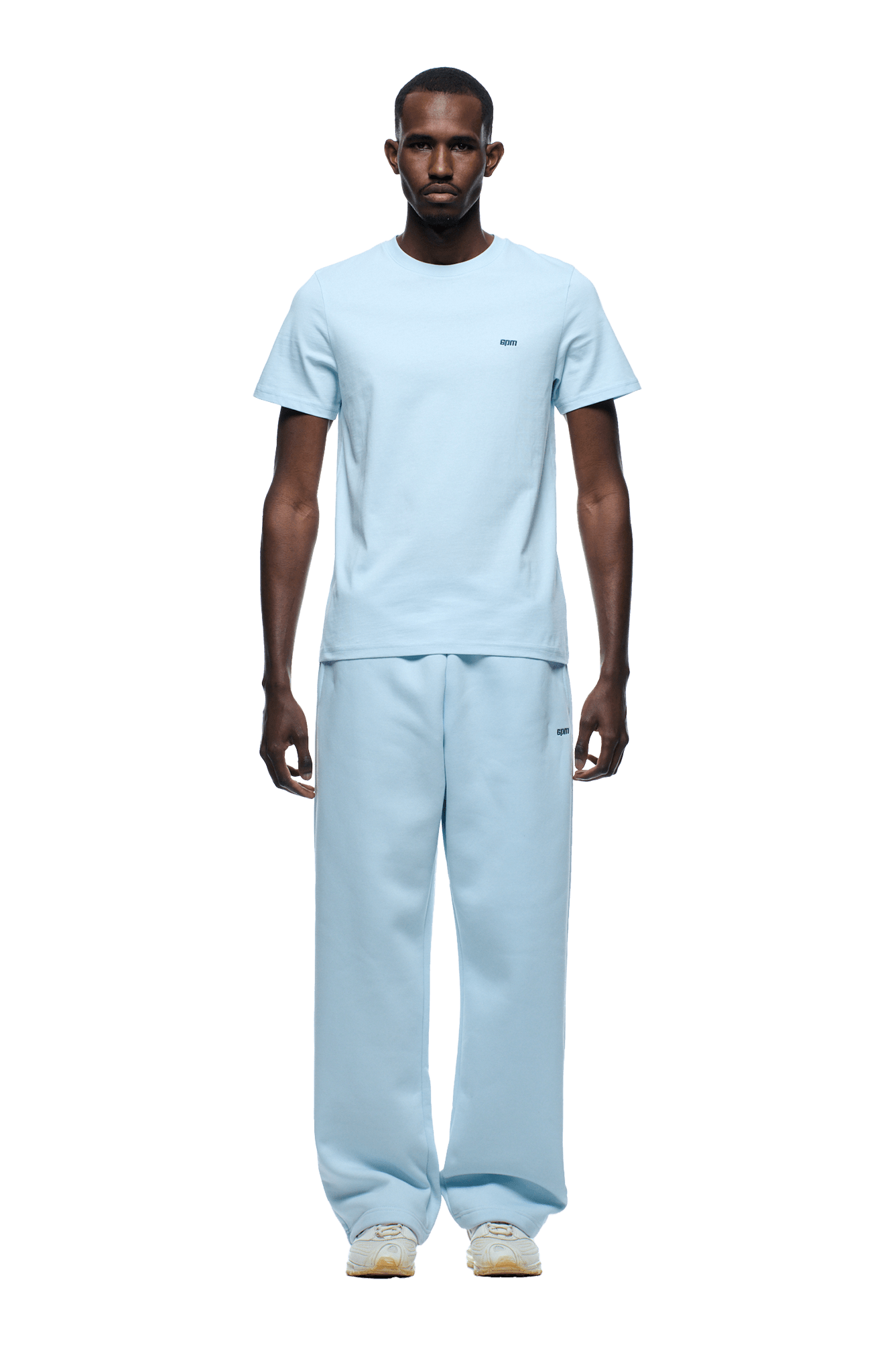 A man faces forward, wearing the 6PM SLIM FIT T-SHIRT ICE BLUE with matching loose sweatpants and white sneakers. The background is transparent.