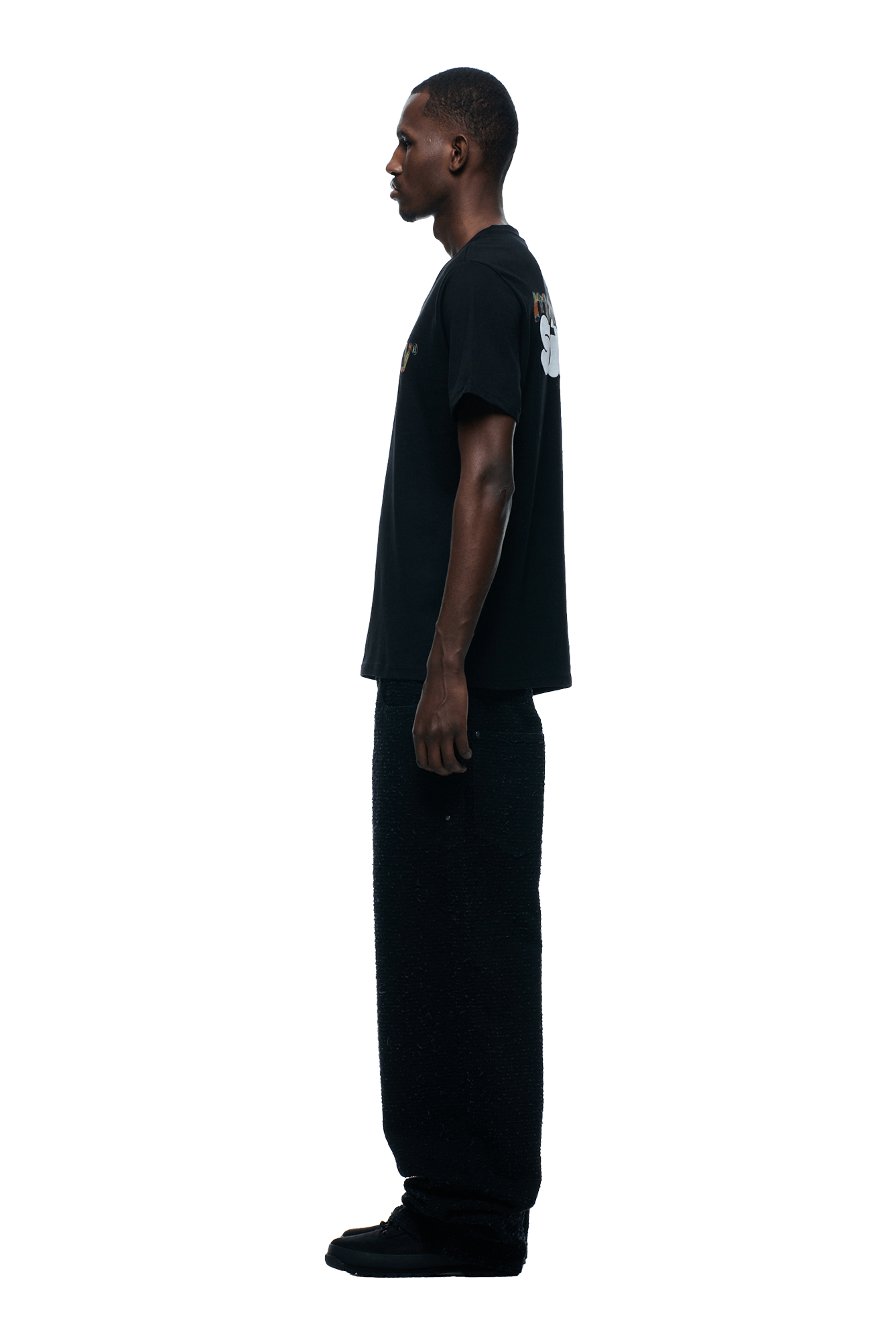 A person in profile faces left, wearing a 6PM TRUE STORY T-SHIRT BLACK with wide black pants and black shoes, set against a plain green background.