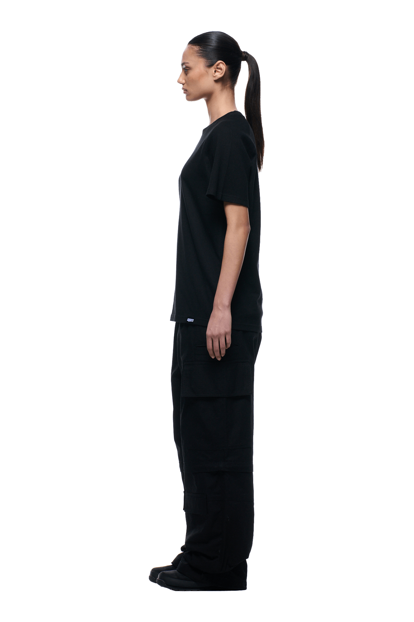 A woman with a ponytail in profile faces left, wearing the 6PM WAFFLE SLEEVE RAGLAN TEE BLACK with black cargo pants and black shoes against a plain background.