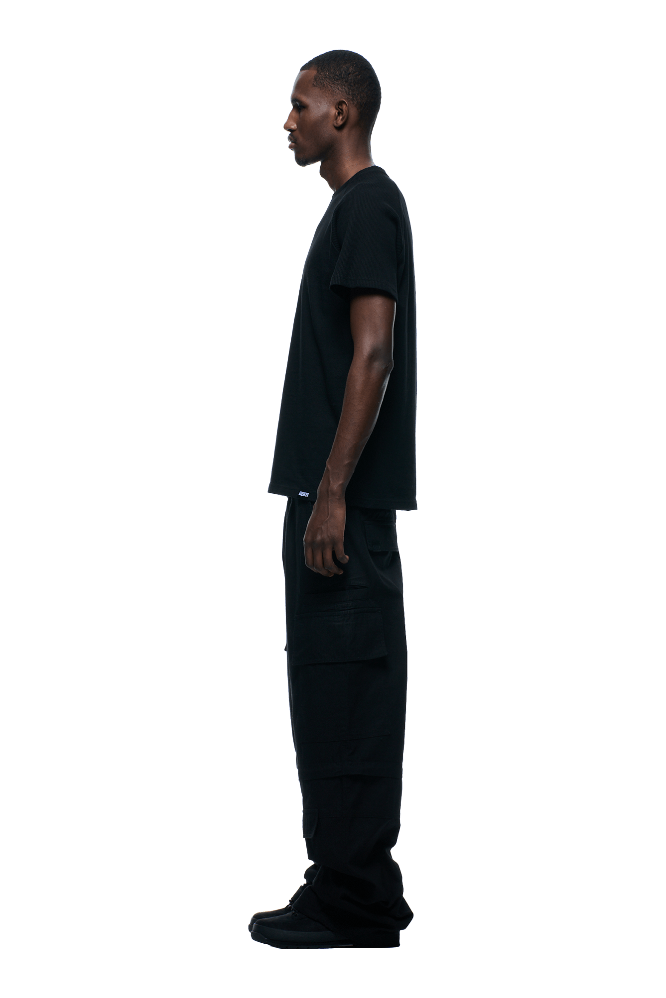 A man in profile faces left, wearing the 6PM WAFFLE SLEEVE RAGLAN TEE BLACK, black cargo pants, and black shoes against a plain transparent background.