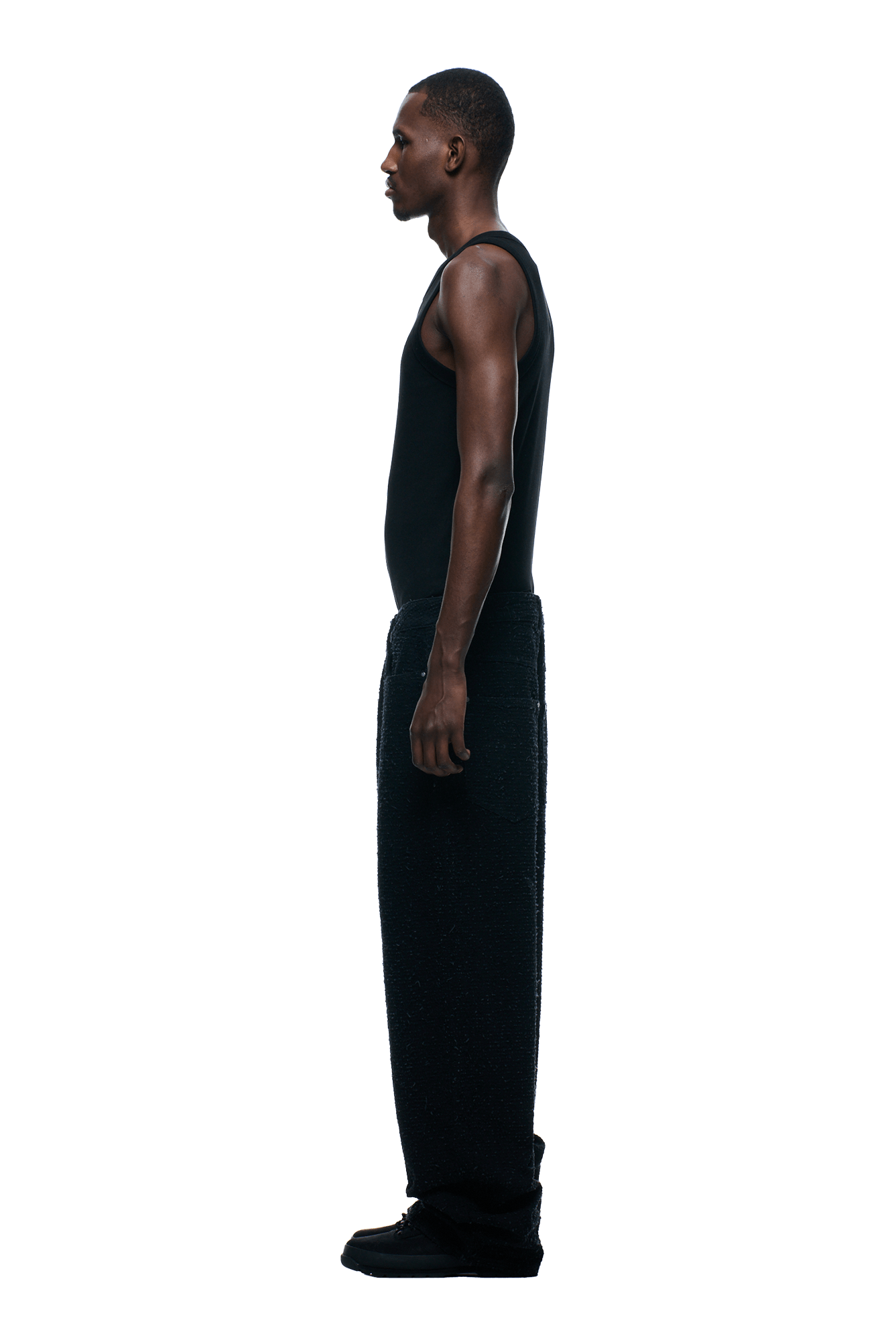 A person in profile faces left, wearing 6PMs NEEDLEPUNCH DENIM X-TRA WIDE pants with a black sleeveless top and black shoes, set against a plain background.