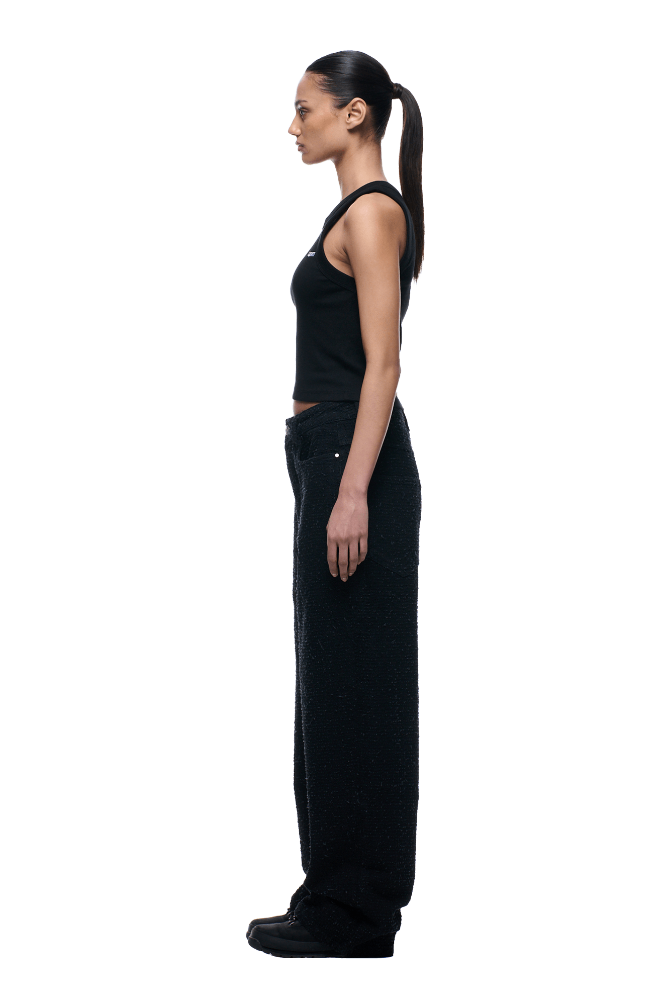A woman stands in profile facing left, wearing 6PMs NEEDLEPUNCH DENIM X-TRA WIDE pants with a black tank top and shoes. Her hair is in a low ponytail. The background is transparent.