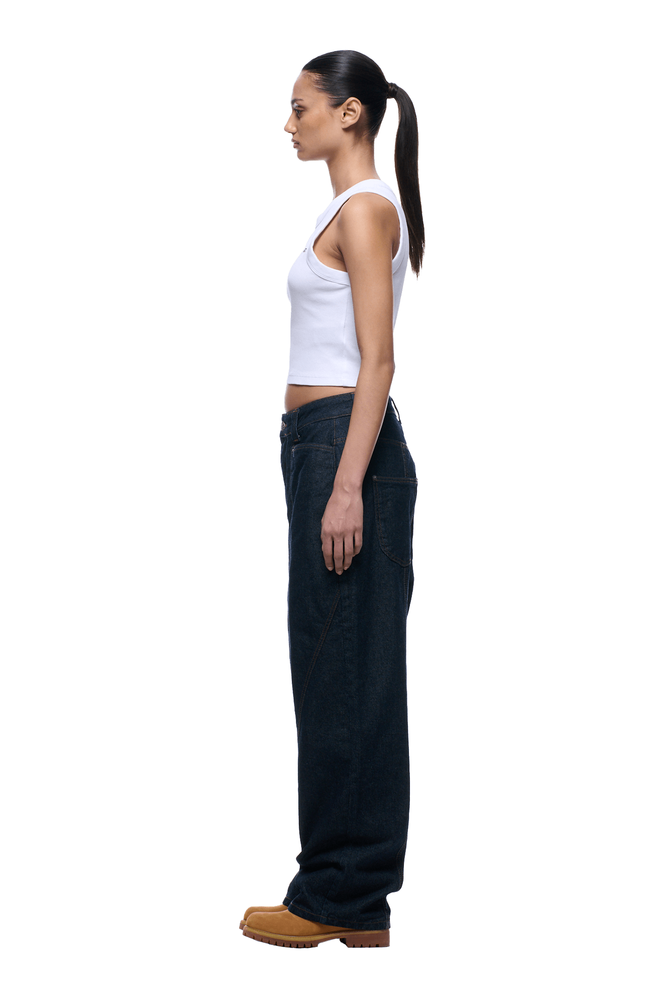 A woman with a ponytail stands in profile against a plain background, wearing a white tank top, tan shoes, and 6PM TYPE 6 DENIM BLUE RAW jeans.