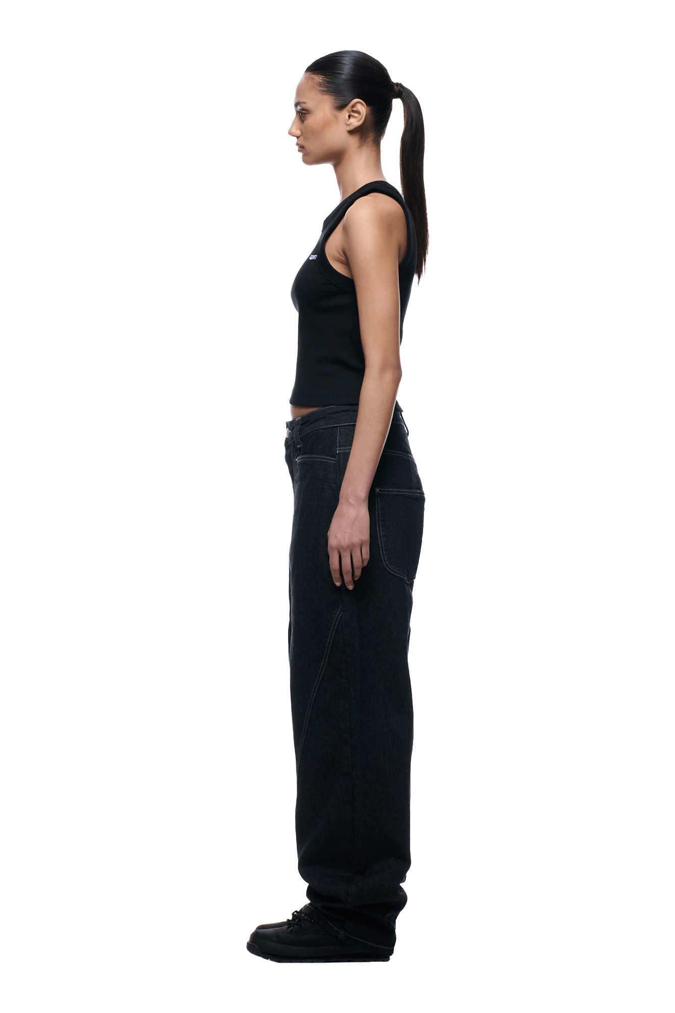 A woman with a long ponytail stands in profile, wearing 6PMs TYPE 6 DENIM BLACK RAW jeans, a black sleeveless top, and black shoes against a plain transparent background.