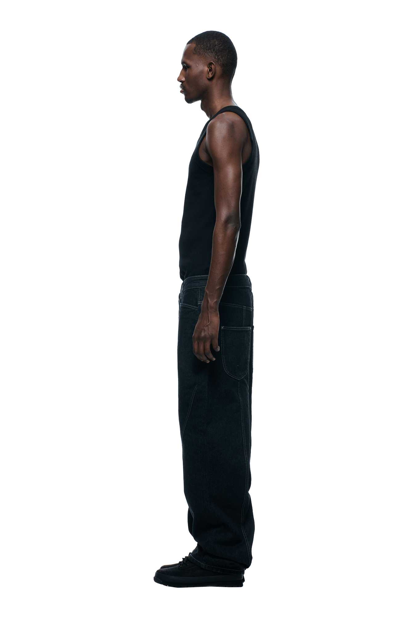 A man stands in profile facing left, wearing a black tank top, black shoes, and 6PM TYPE 6 DENIM BLACK RAW jeans against a plain background.
