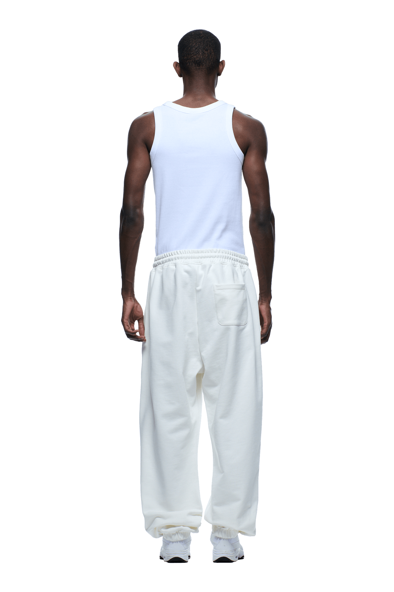 A man faces away, wearing a 6PM SUPER WIDE JOGGER COCONUT WHITE sweatpants, a white sleeveless tank top, and white sneakers against a transparent background.