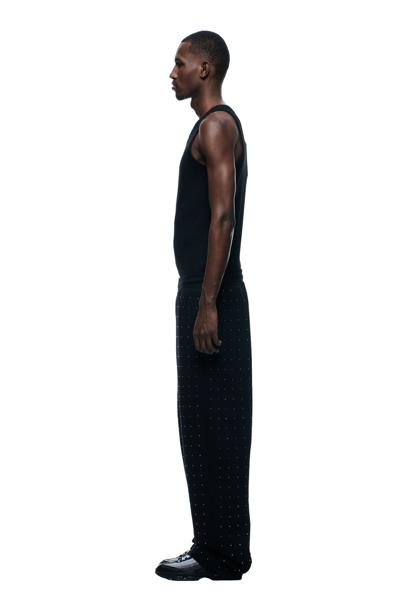 A man stands in profile facing left, wearing 6PMs RHINESTONE OPEN LEG JOGGER BLACK pants with a dotted pattern, a black sleeveless shirt, and black shoes against a plain background.