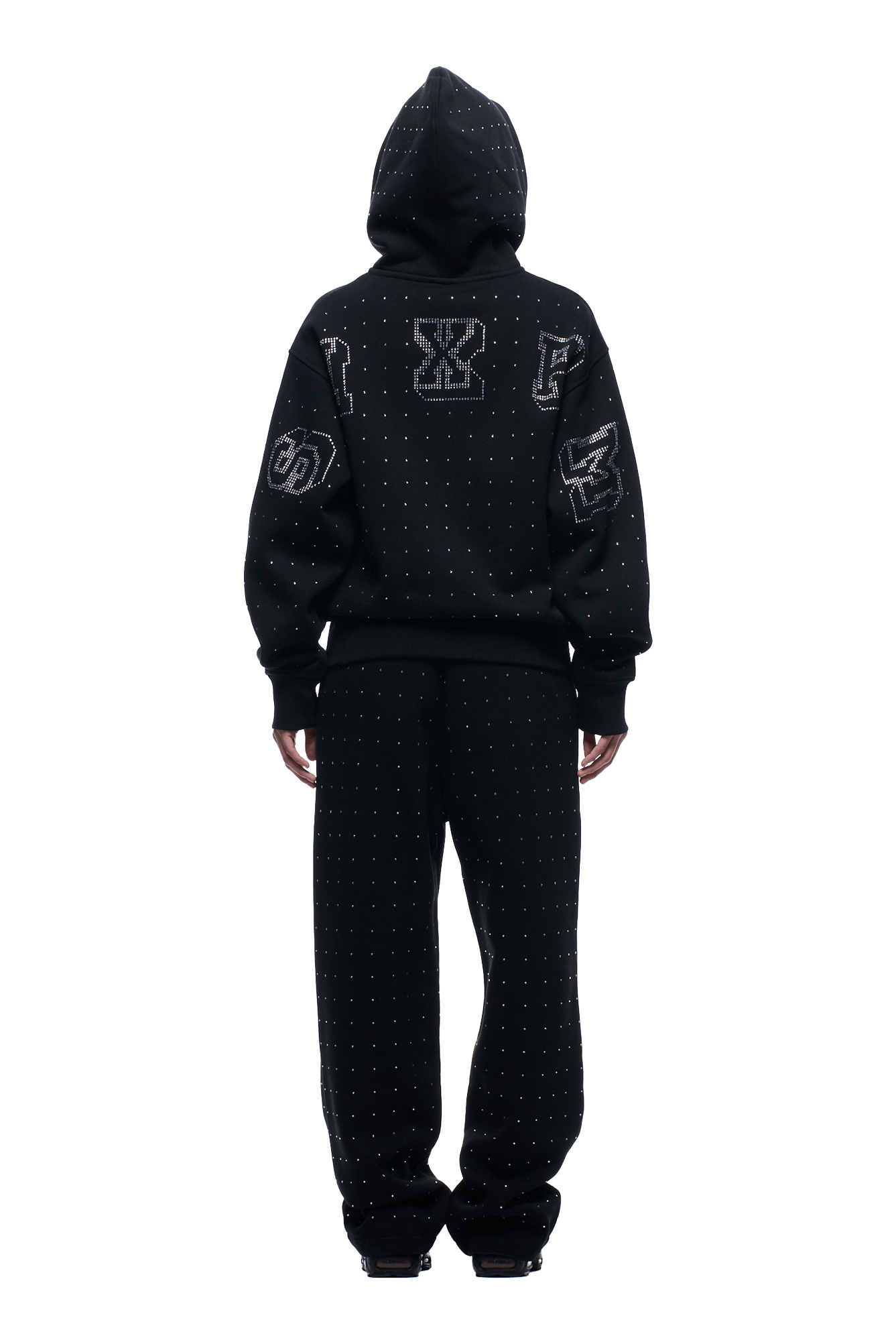A person faces away wearing the 6PM RHINESTONE LETTER ZIP HOODIE BLACK, a loose-fit black hoodie with rhinestone letter details and large graphic patches on the back and sleeves. The hood is up, completing the stylish look.