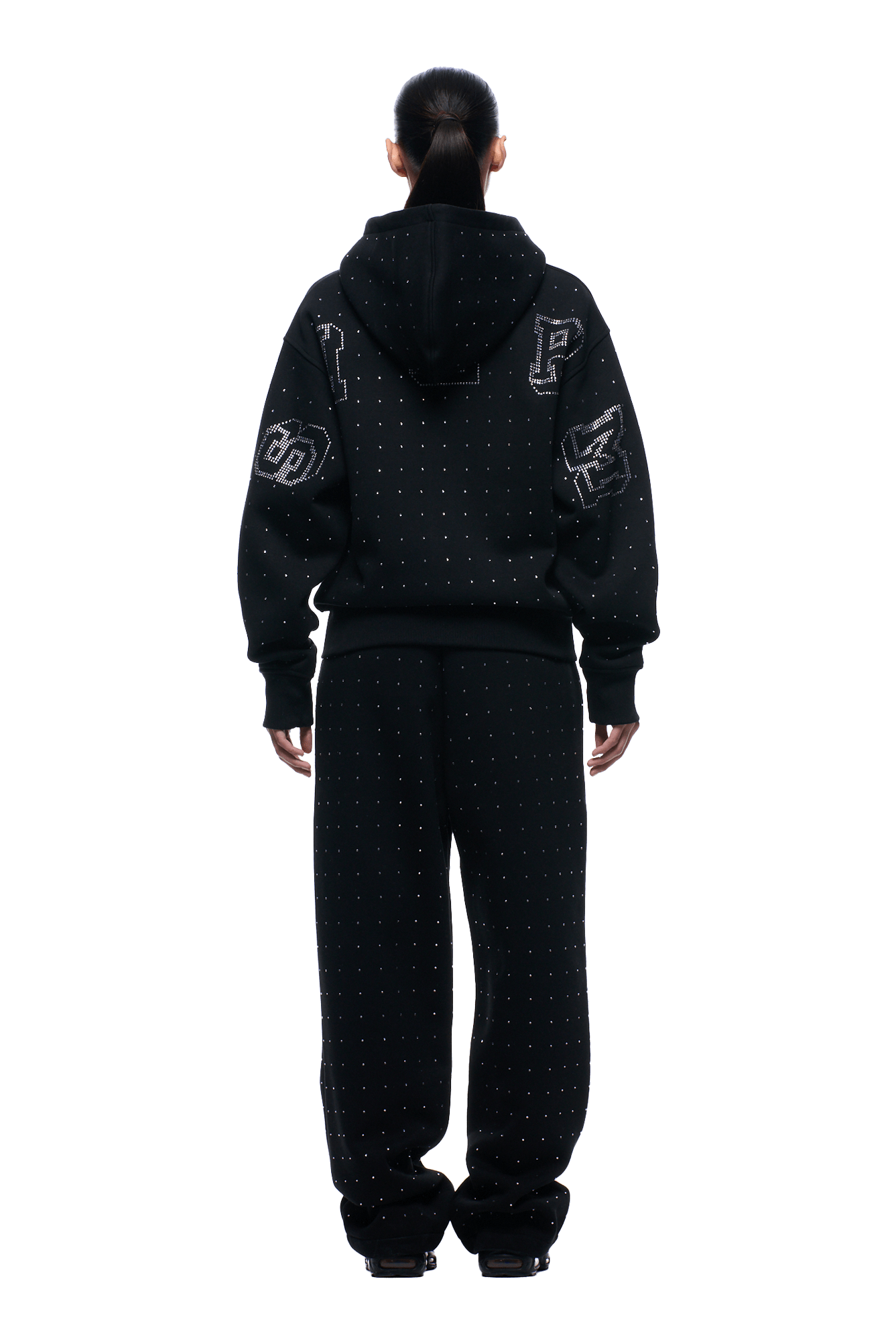 A person stands facing away against a plain background, wearing the 6PM RHINESTONE LETTER ZIP HOODIE BLACK featuring small dotted patterns and large patch designs on the sleeves and back, paired with matching sweatpants.
