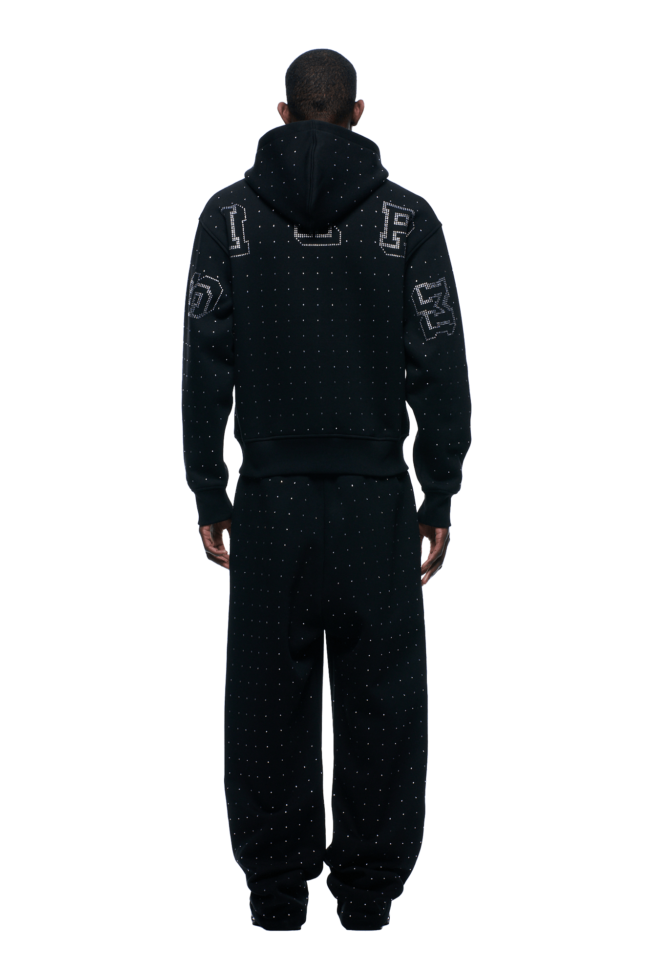 A person stands facing away, wearing the 6PM RHINESTONE LETTER ZIP HOODIE BLACK with bold sleeve and hood patches and small white dotted details, paired with matching baggy pants, against a plain background.