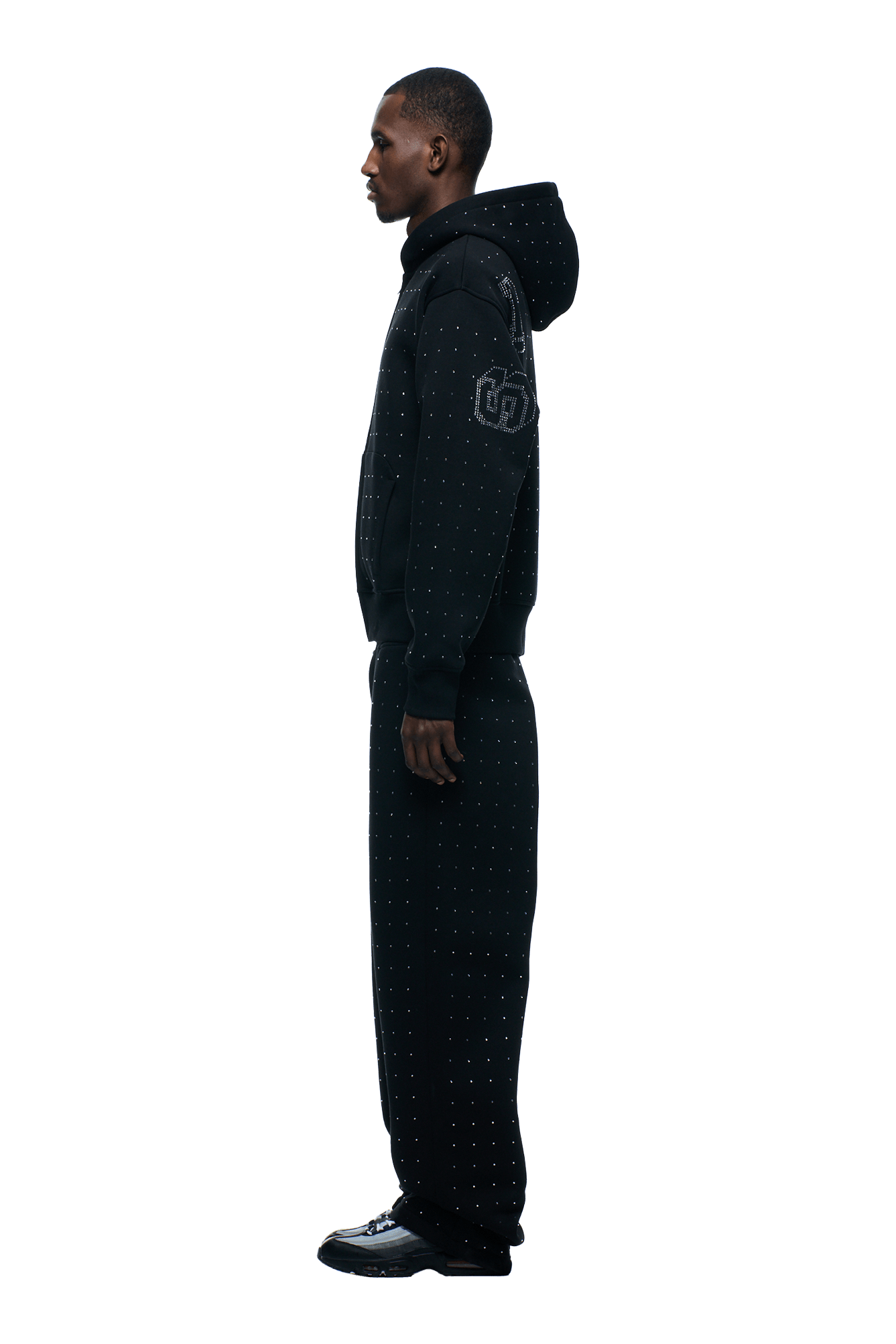 A man stands in profile wearing the 6PM RHINESTONE LETTER ZIP HOODIE BLACK and matching pants, paired with black shoes. The background is transparent.