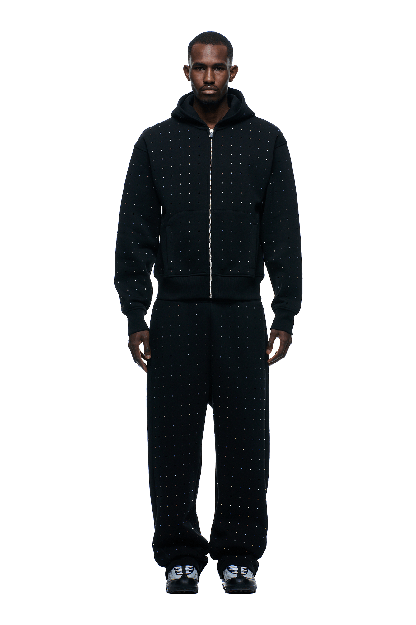A man faces forward against a plain green background, wearing the 6PM RHINESTONE LETTER ZIP HOODIE BLACK with matching black pants featuring a subtle dotted pattern and black sneakers.