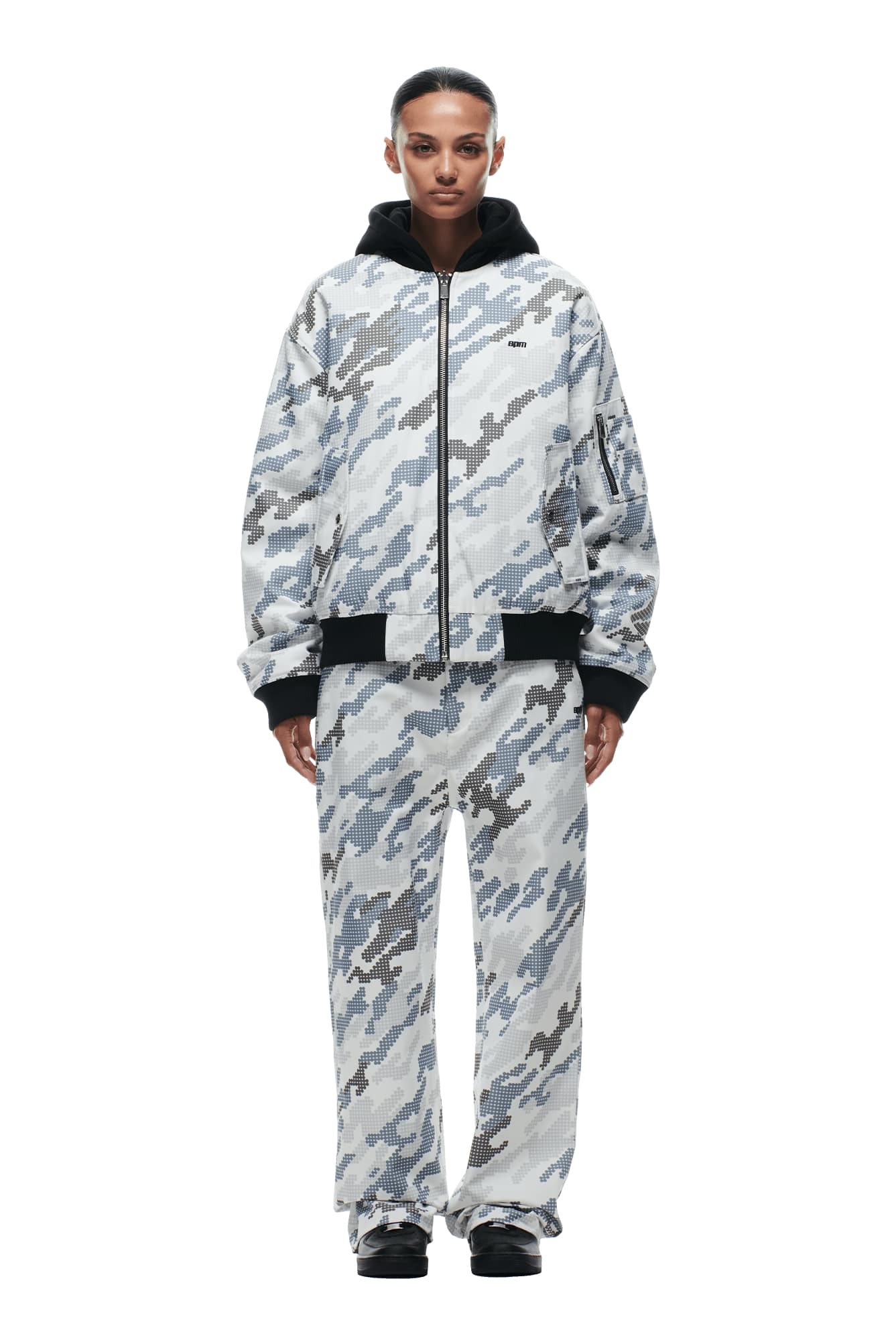 A person stands facing forward against a white background, wearing the 6PM DOT CAMO HOODED BOMBER JACKET in light gray and white camouflage with black accents and a black hood.