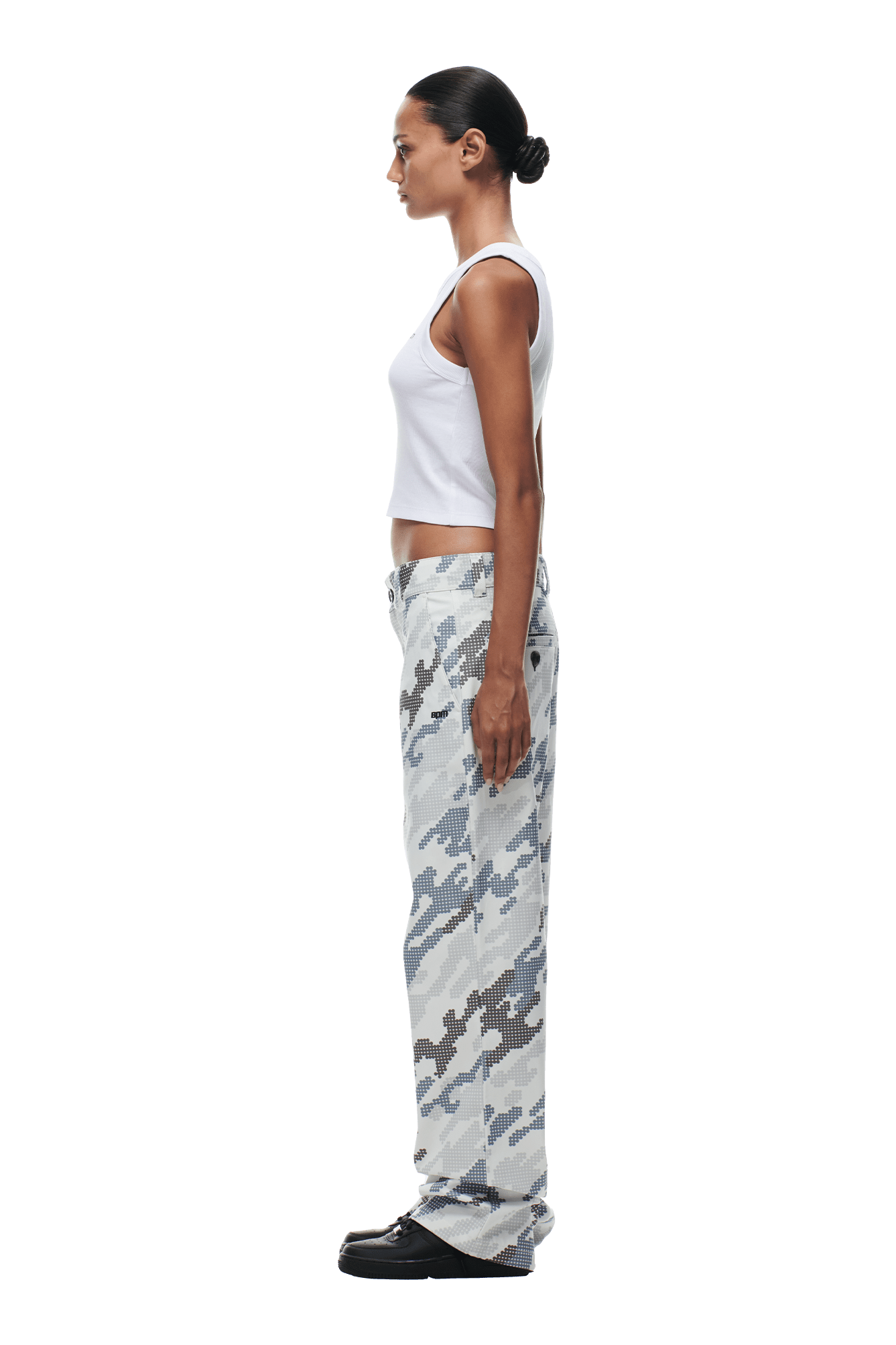 A woman stands in profile against a plain background, wearing 6PM’s DOT CAMO CHINO PANTS with a white tank top and black shoes. Her hair is styled in a neat bun.