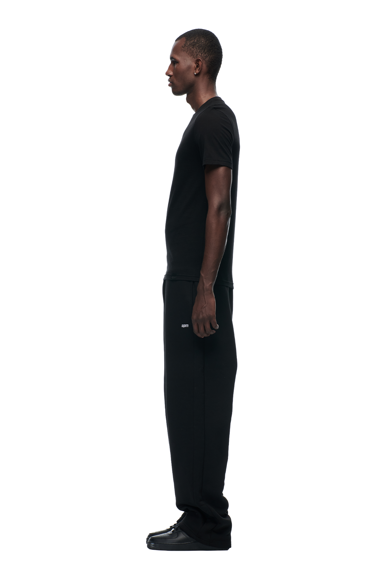 A man stands in profile facing left, wearing the 6PM SLIM FIT T-SHIRT BLACK with black pants and shoes, against a plain white background.