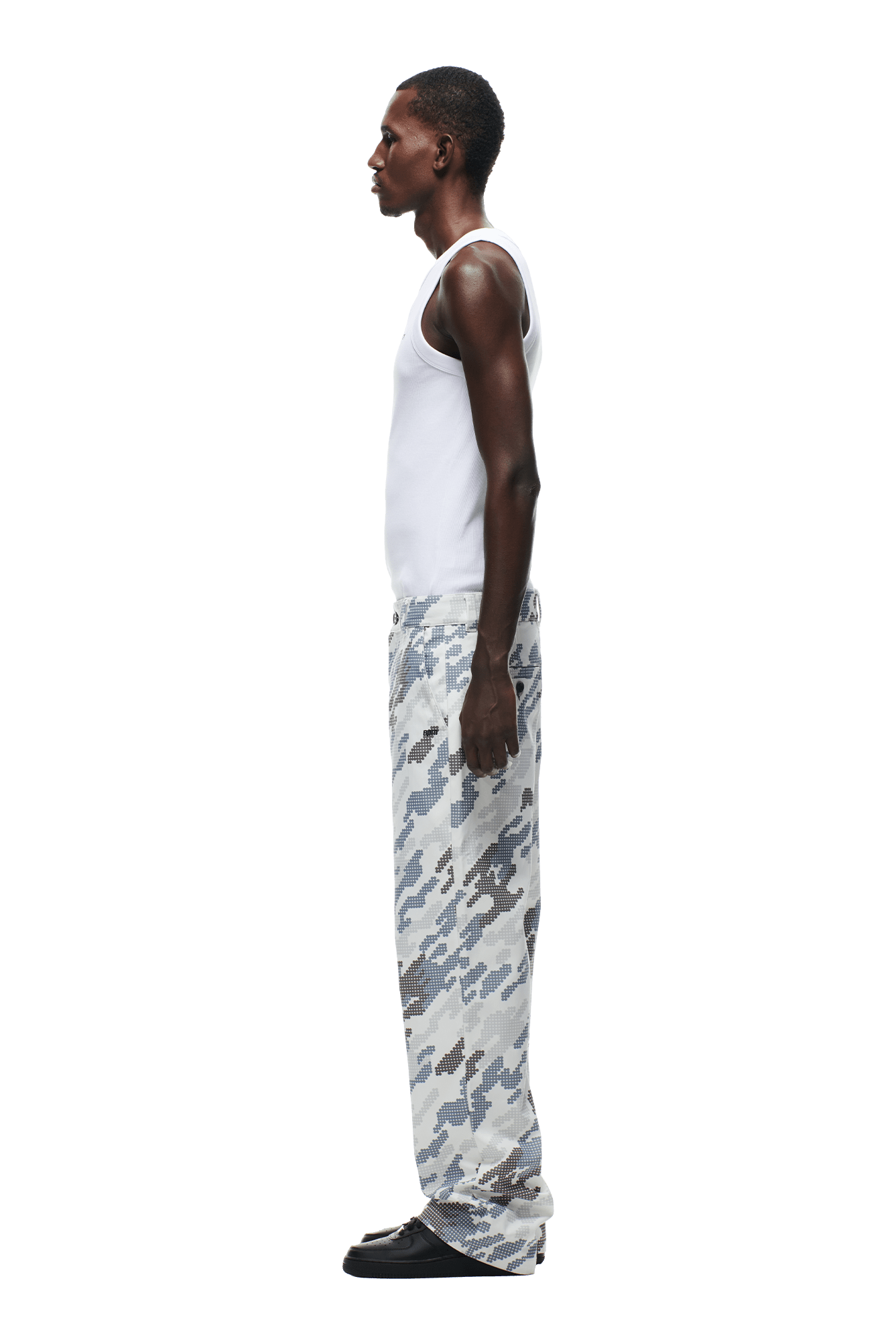 A man in profile, facing left, wears a white tank top, 6PM DOT CAMO CHINO PANTS in gray and white, and black shoes against a plain green background.