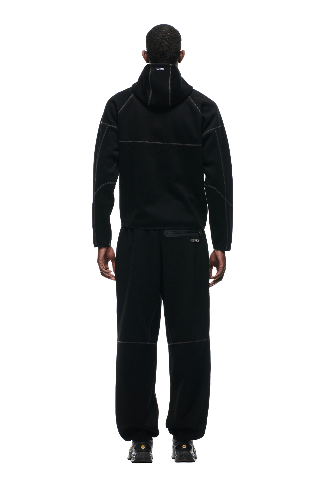 A person faces away from the camera, wearing the 6PM HOODED ROADMAN JACKET in black with subtle white stitching, paired with black pants and shoes. The background is transparent.