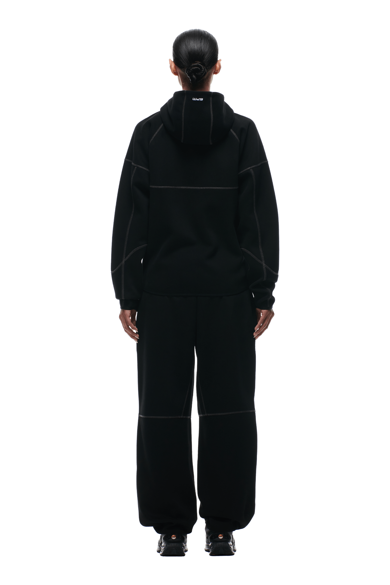 A person with dark hair in a low bun stands with their back to the camera, wearing the 6PM HOODED ROADMAN JACKET and matching black pants with visible seams and black shoes, against a green background.