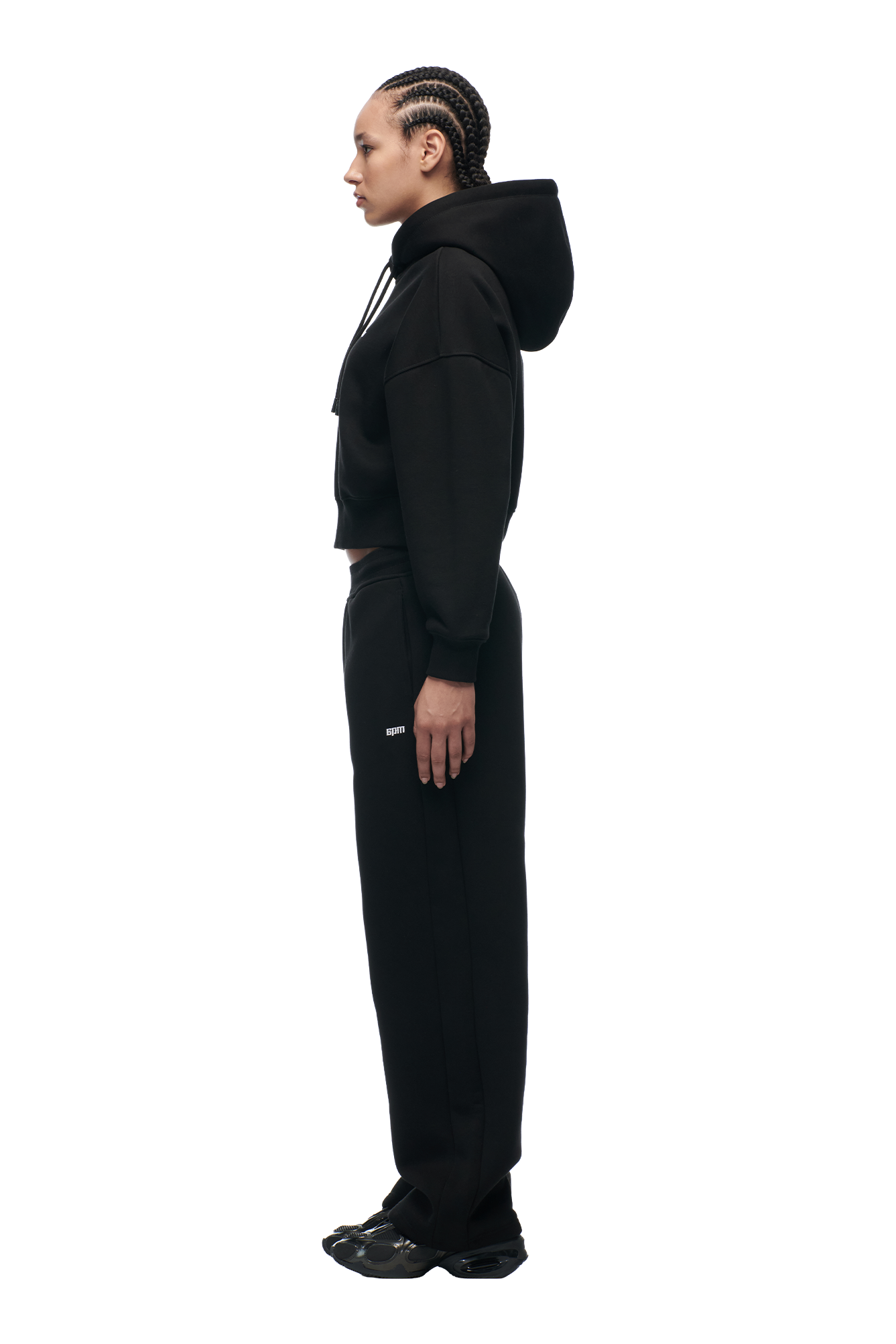 A person with braided hair stands in profile wearing the 6PM WMNS CROPPED HOODIE BLACK, paired with black sweatpants and sneakers, against a plain white background.
