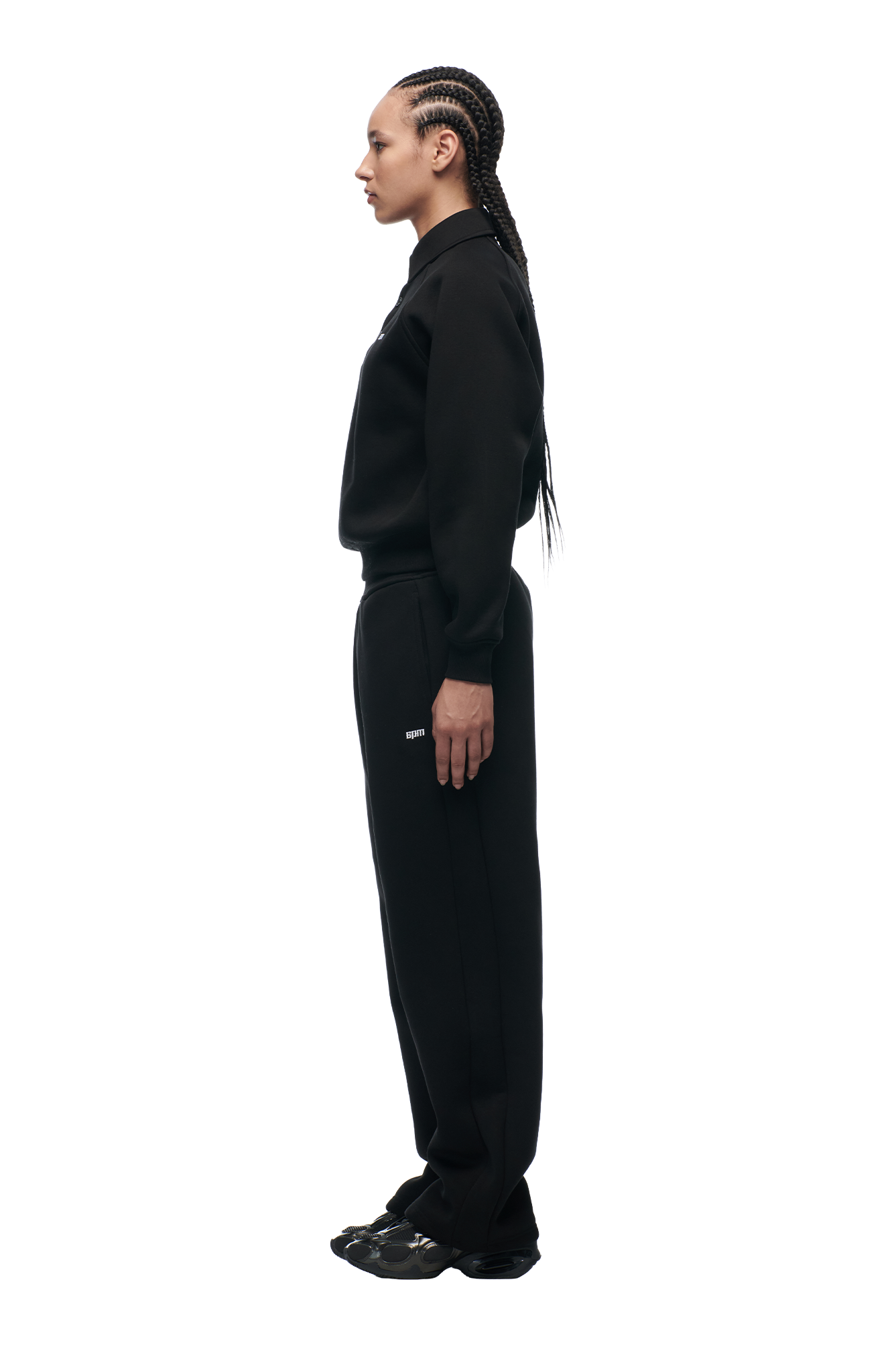 A person with long braids stands in profile, facing left, wearing the 6PM WMNS POLO BLACK sweatshirt with matching black pants and black sneakers against a plain white background.