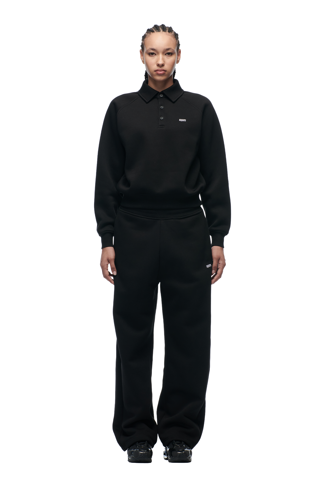 A person stands facing forward, wearing the 6PM WMNS POLO BLACK collared sweatshirt and loose black pants with black shoes against a plain white background.