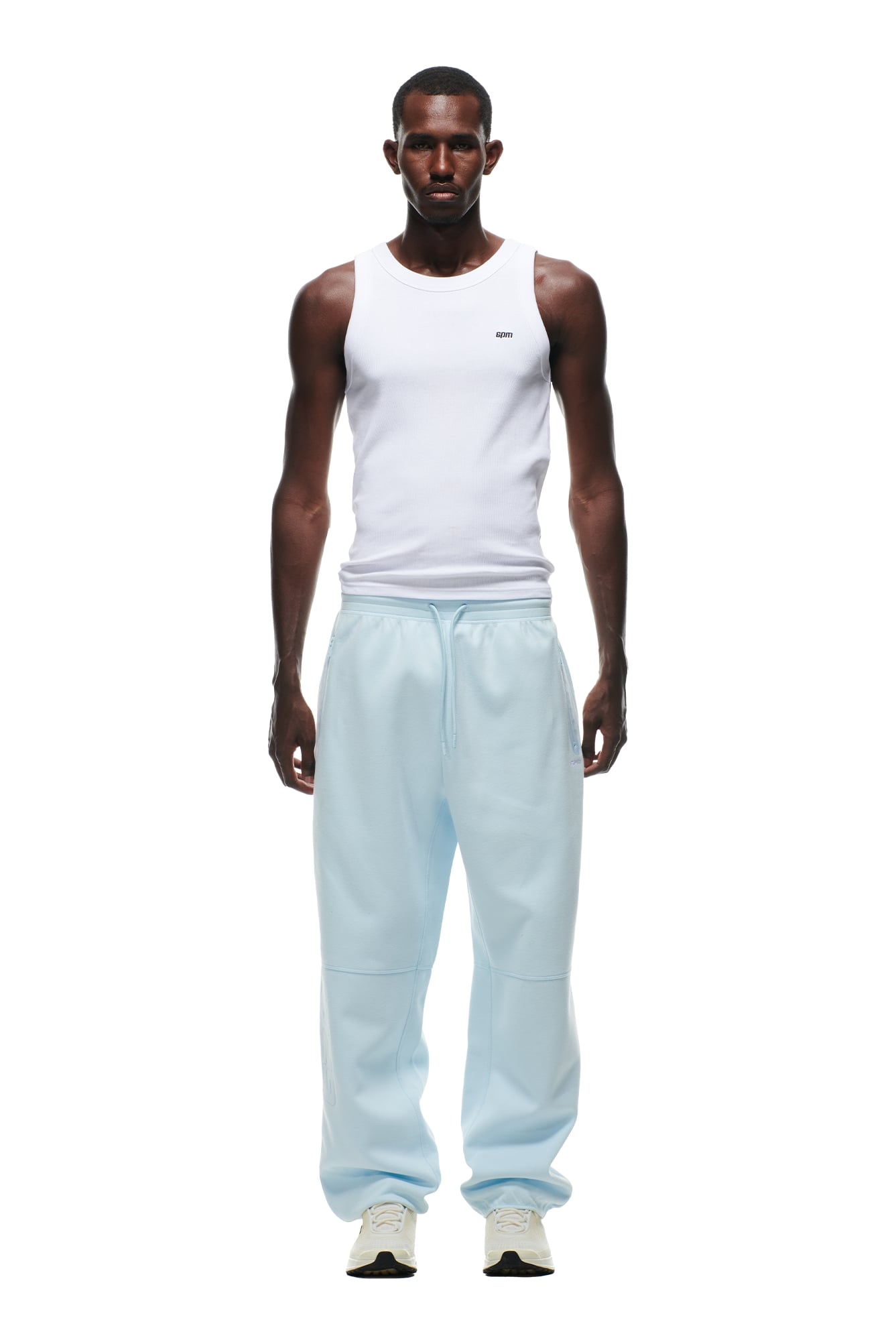 A man faces forward, wearing a white sleeveless tank top, 6PM WIDE ROADMAN PANTS ICE BLUE sweatpants, and white sneakers against a plain white background.