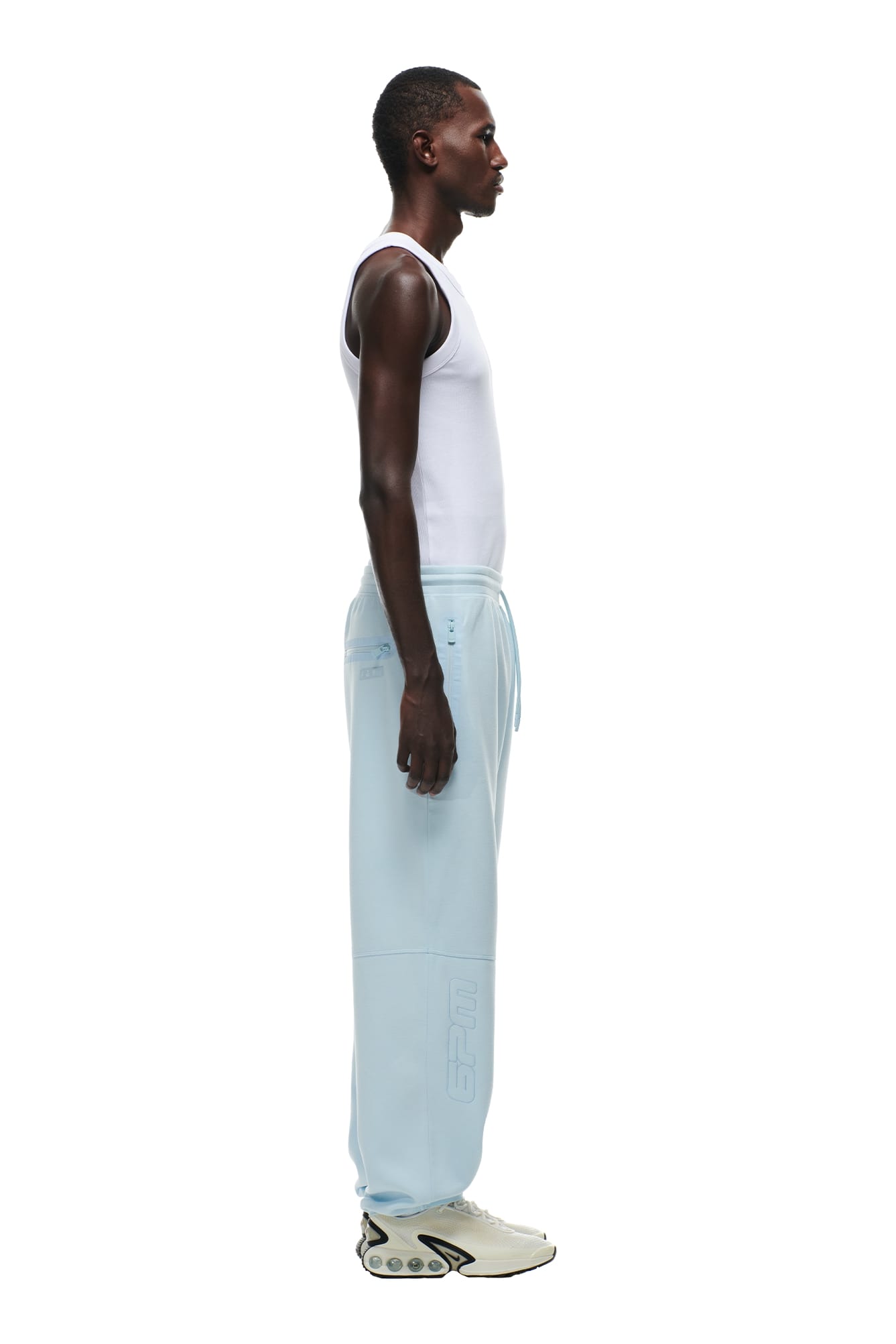 A person stands in profile against a white background, wearing a white sleeveless shirt, 6PM WIDE ROADMAN PANTS ICE BLUE with lettering on the leg, and beige sneakers featuring a unique sole design.