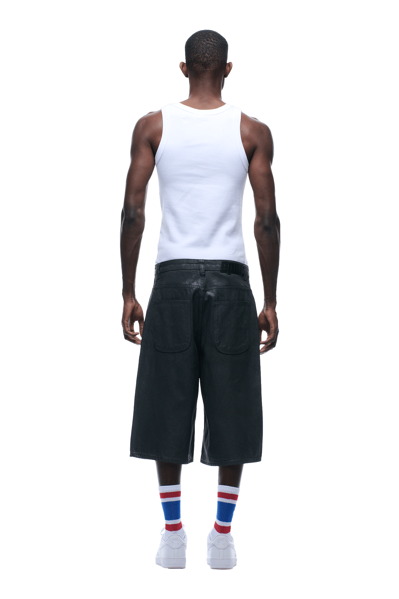 A person stands facing away, wearing COATED DENIM SHORTS DARK CHROME with a white sleeveless shirt, white striped socks, and white sneakers against a plain background.