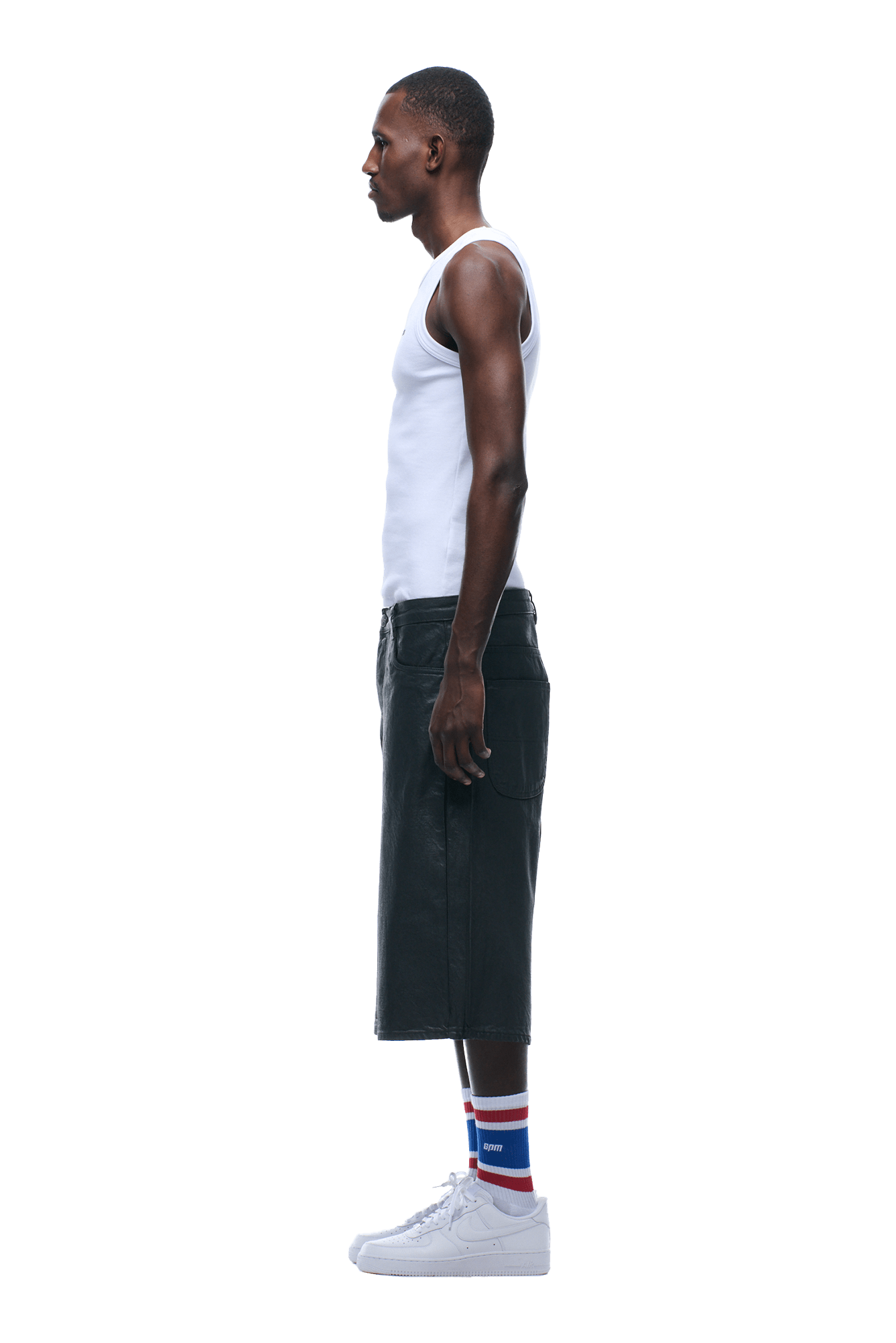 A man stands in profile, wearing a white tank top, COATED DENIM SHORTS DARK CHROME, white sneakers, and white socks with red and blue stripes. The background is transparent.