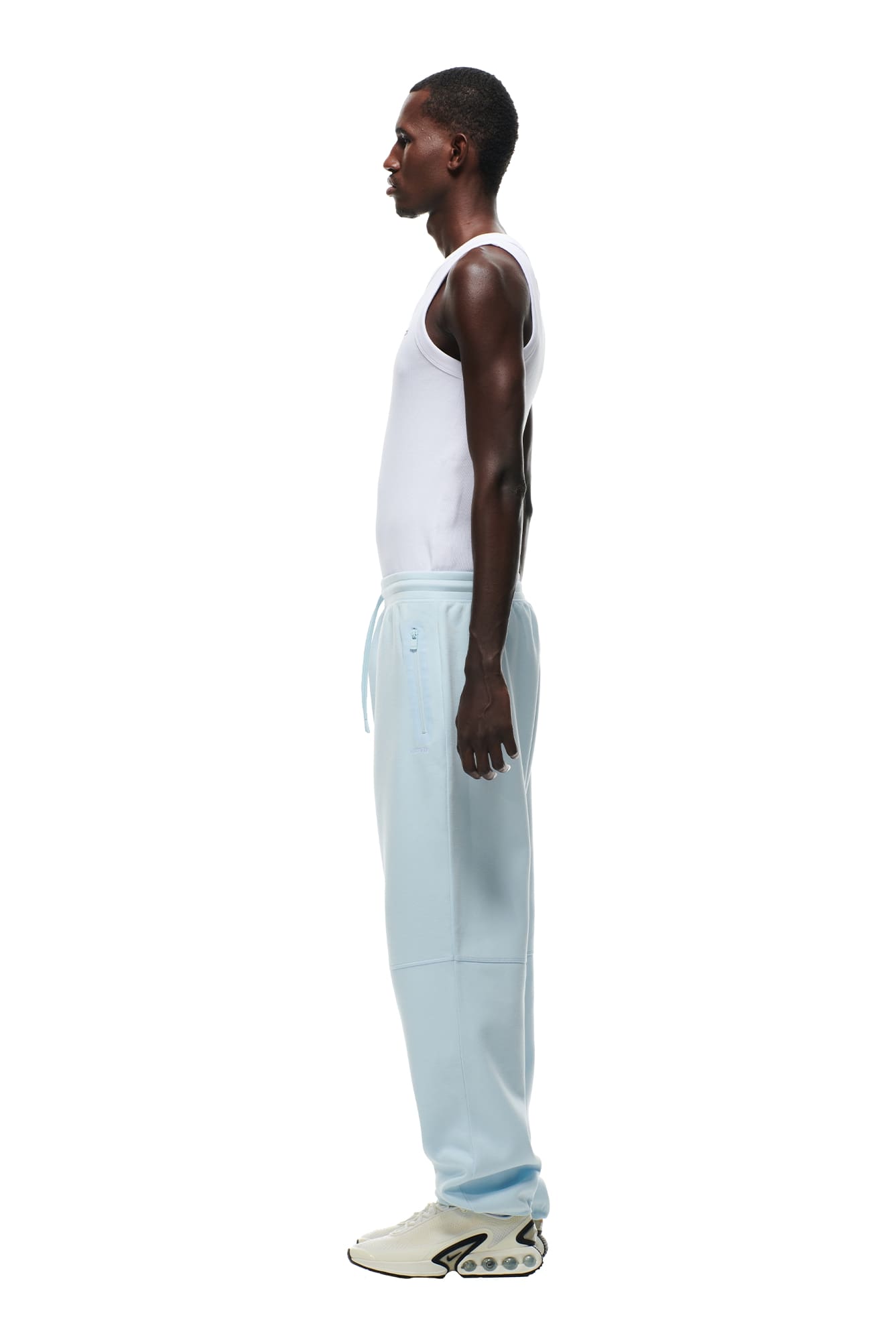 A man stands in profile against a white background, wearing a white tank top, 6PM WIDE ROADMAN PANTS in ice blue, and white sneakers with a unique sole design.