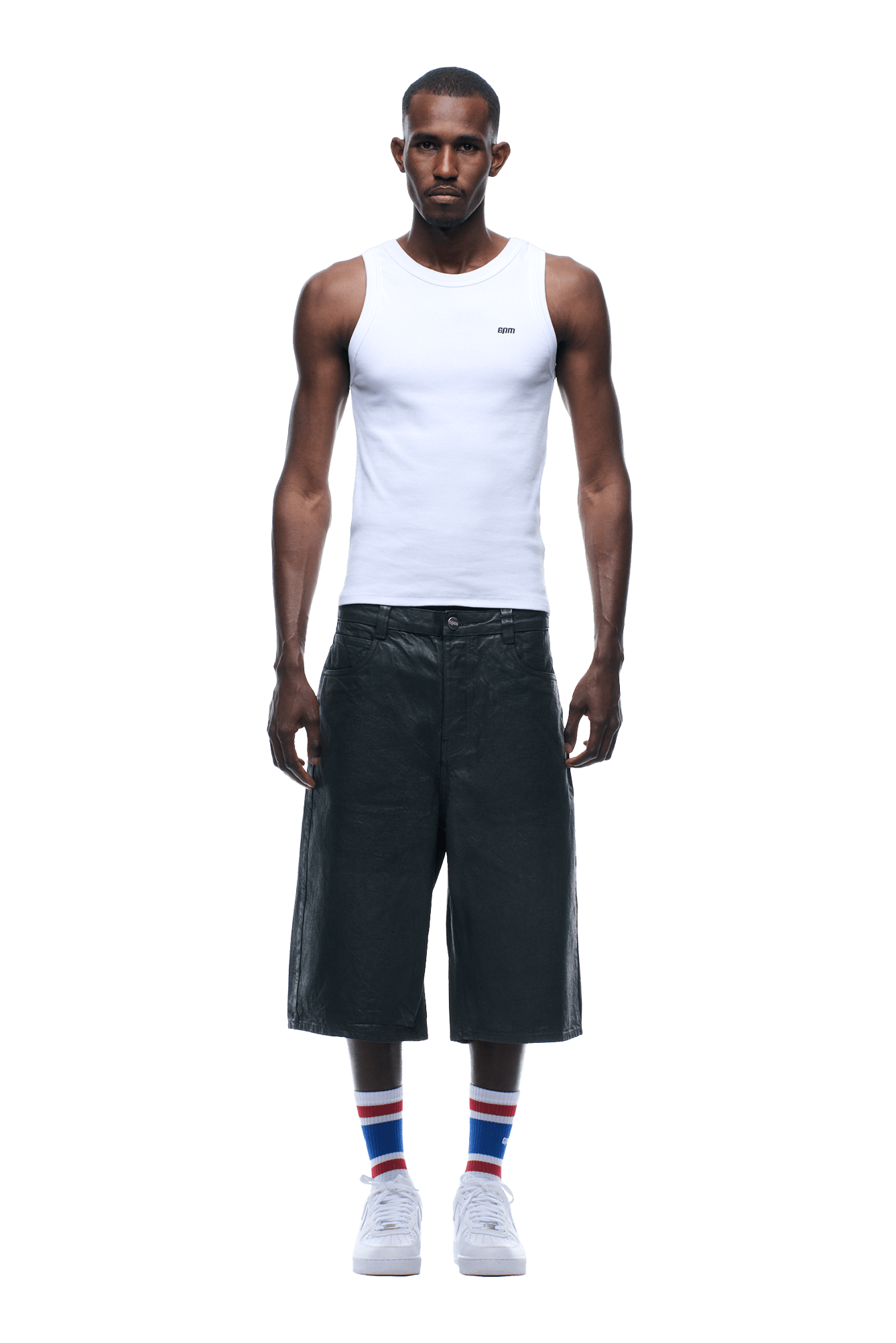 A man faces forward, wearing COATED DENIM SHORTS DARK CHROME, a white sleeveless tank top, white sneakers, and white socks with blue and red stripes. The background is transparent.