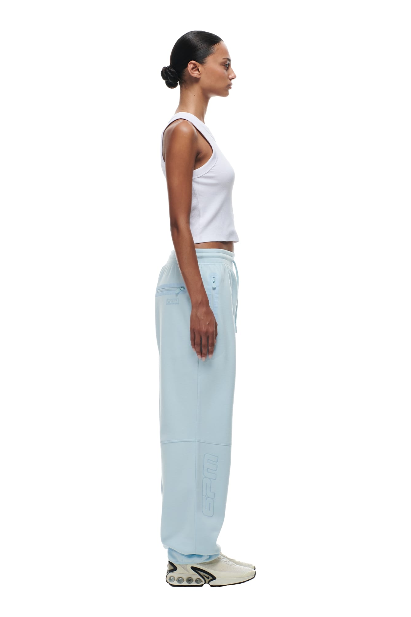 A woman stands in profile against a white background, wearing a white tank top, 6PM WIDE ROADMAN PANTS ICE BLUE with drawstrings and subtle graphics, cream sneakers with a unique sole design, and her hair pulled back in a neat bun.