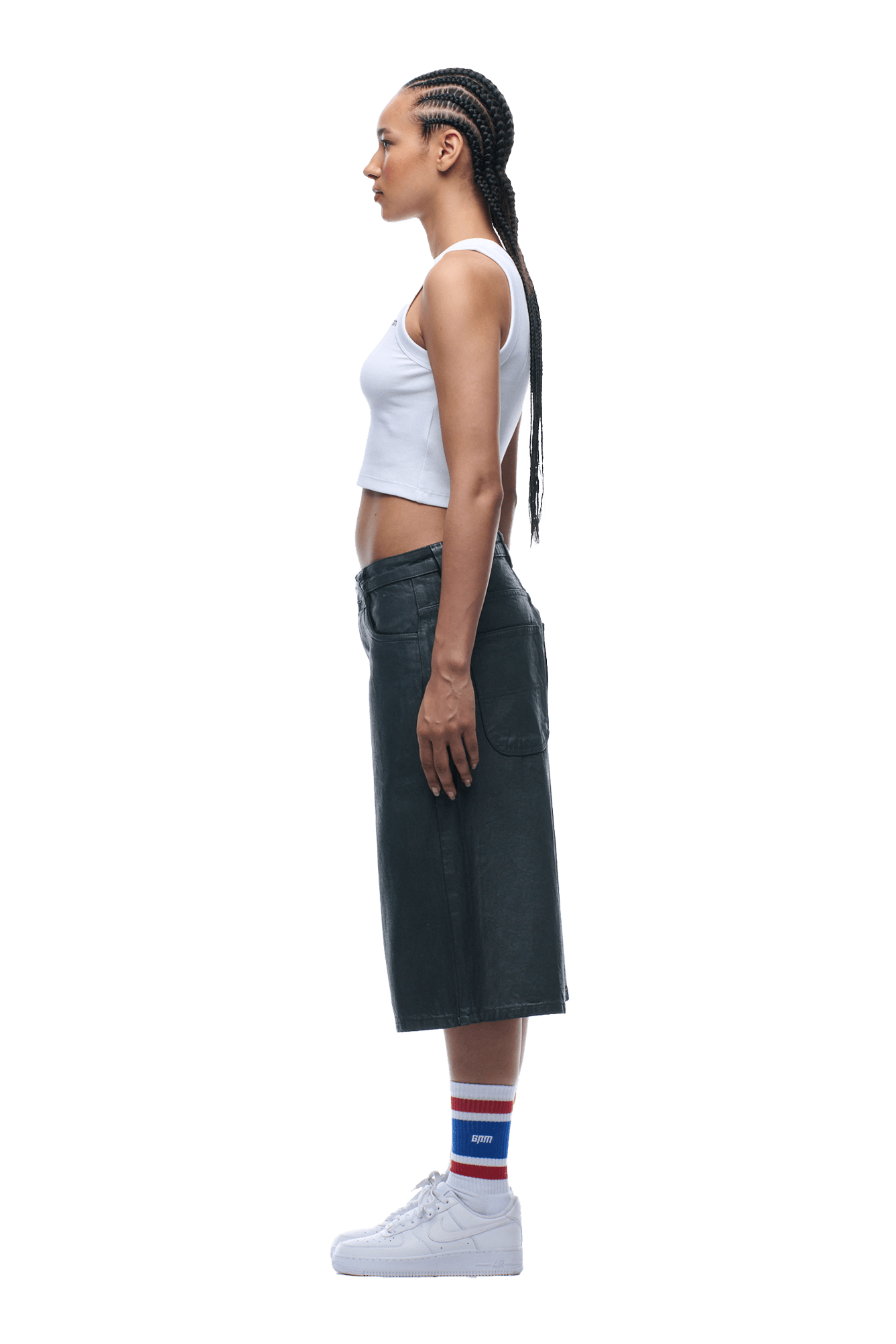 A woman with long braided hair stands in profile against a white background, wearing COATED DENIM SHORTS DARK CHROME, a white crop top, striped socks, and white sneakers.