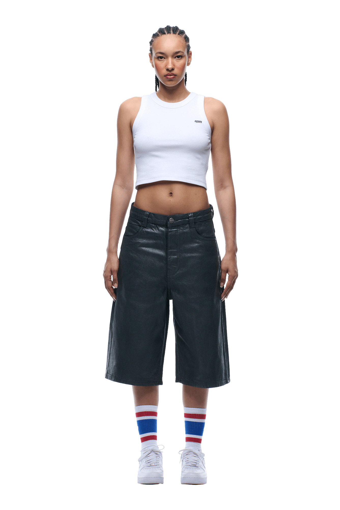 A person faces forward, wearing a white sleeveless crop top, COATED DENIM SHORTS DARK CHROME, white sneakers, and white socks with blue and red stripes. Their hair is in cornrows. The background is transparent.