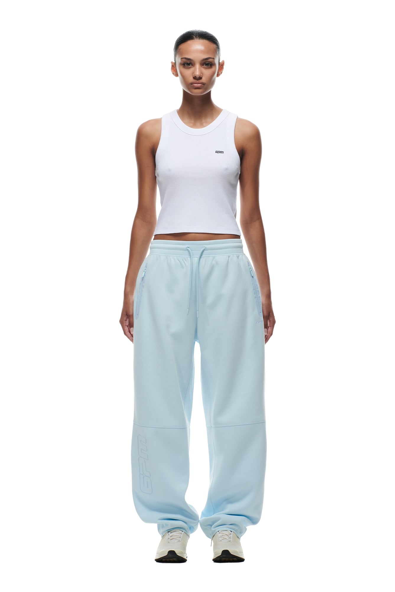 A woman stands facing forward in 6PMs WIDE ROADMAN PANTS ICE BLUE, a fitted white tank top, and white sneakers. Her hair is pulled back, she has a neutral expression, and the background is plain white.
