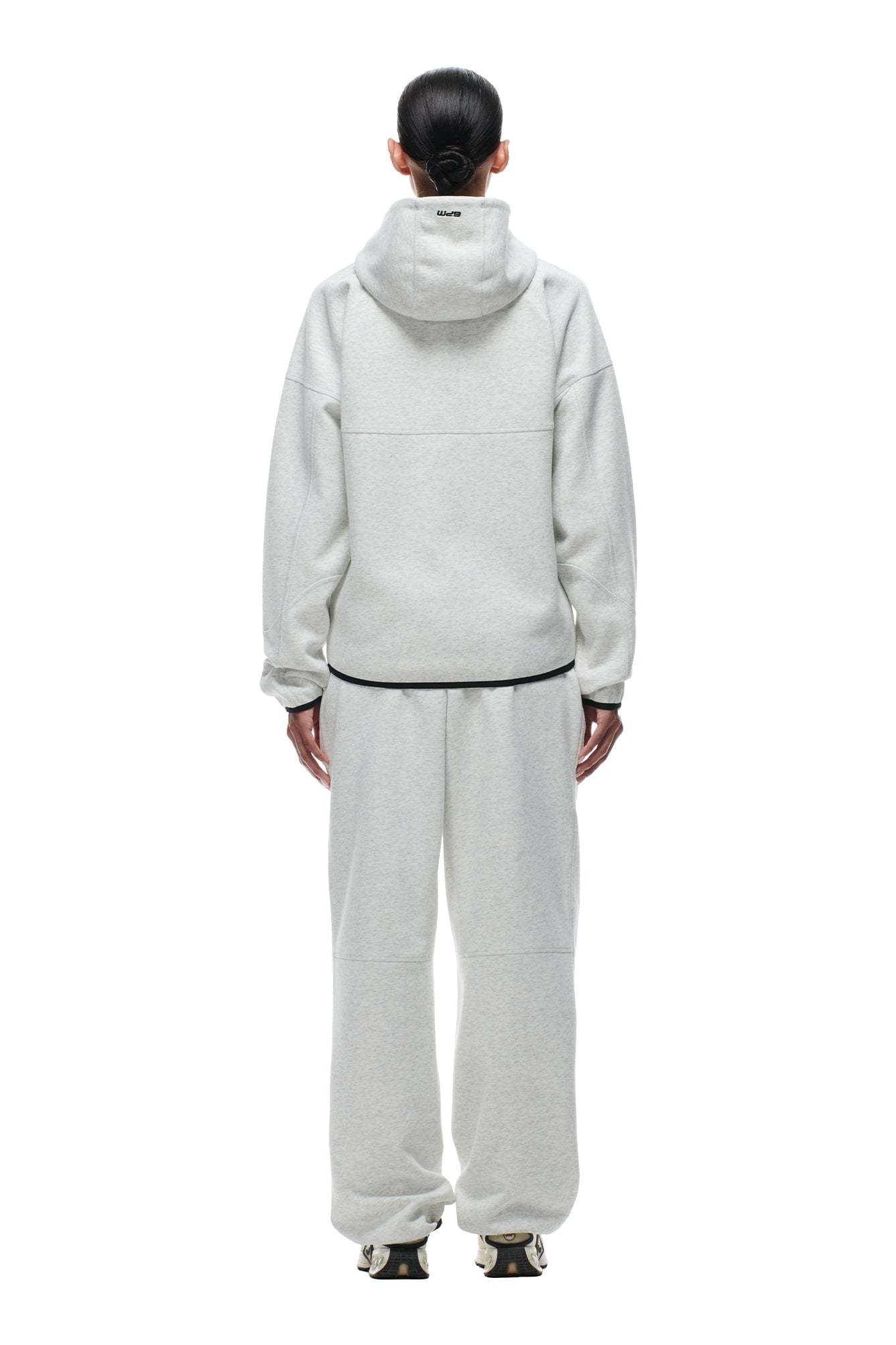 A person faces away in a 6PM HOODED ROADMAN JACKET LIGHT GREY, paired with matching sweatpants and black and white sneakers. Their hair is tied back in a low bun, against a plain white background.