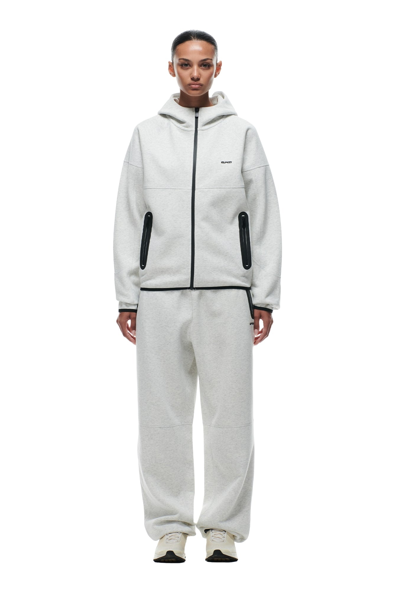 A person stands facing forward, wearing the 6PM HOODED ROADMAN JACKET LIGHT GREY paired with matching sweatpants featuring black trim and white sneakers for a casual, comfortable look.