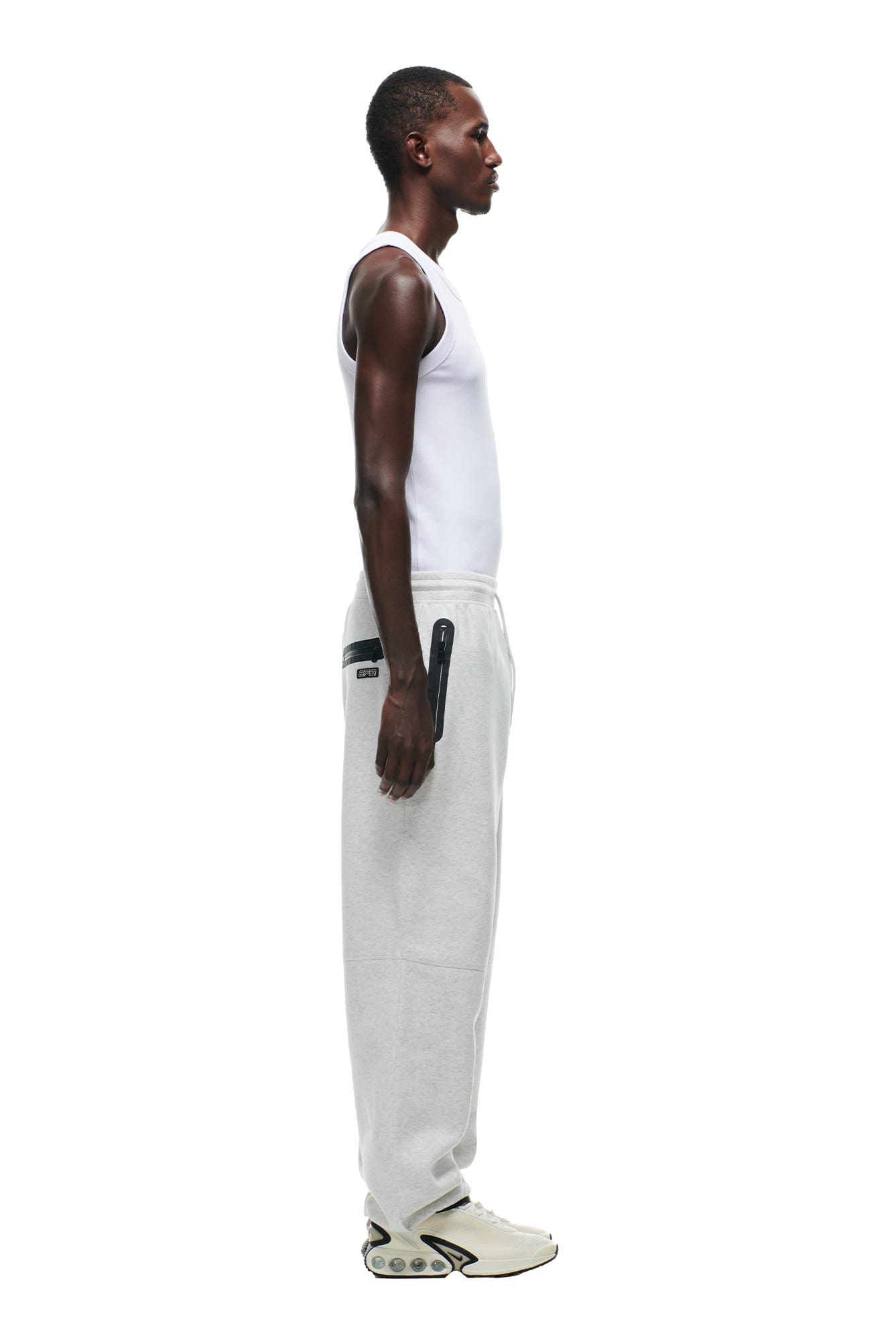 A man in profile wears a white tank top, 6PM WIDE ROADMAN PANTS in light grey melange with side zipper detail, and chunky white sneakers, standing against a plain white background.