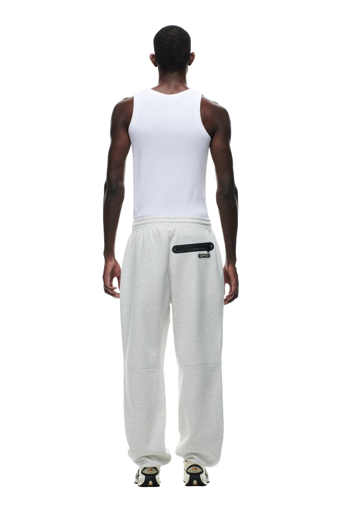 A person faces away from the camera, wearing 6PM WIDE ROADMAN PANTS in light grey melange with a white sleeveless shirt and sneakers, standing against a plain white background.