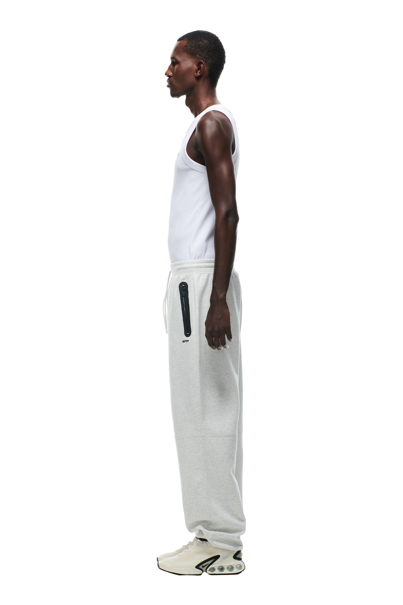 A man in a white tank top, 6PM WIDE ROADMAN PANTS LIGHT GREY MELANGE, and white sneakers stands in profile against a white background.