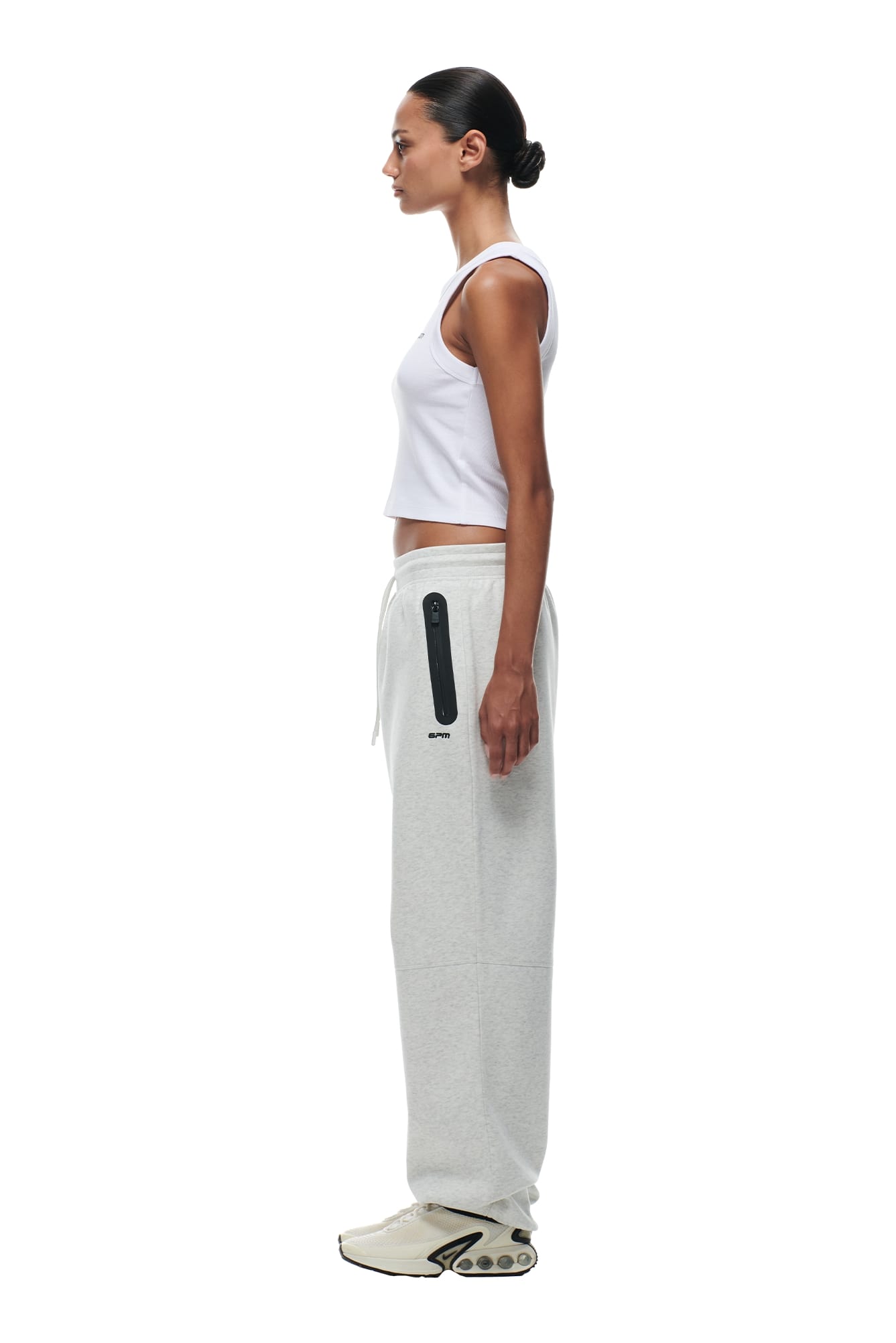 A woman stands in profile against a white background, wearing a white sleeveless crop top, 6PM WIDE ROADMAN PANTS LIGHT GREY MELANGE with a black zipper pocket, and white sneakers. Her hair is tied back in a neat bun.