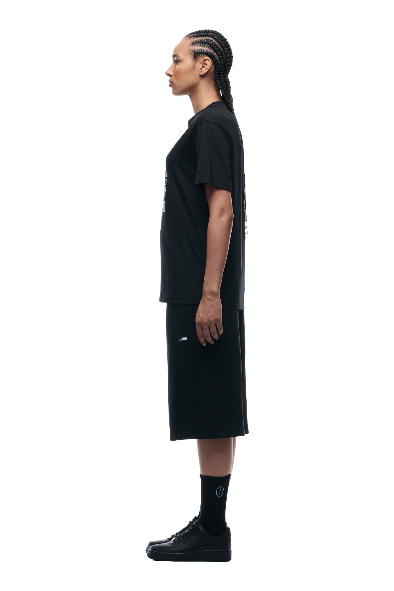A person with braided hair stands in profile, facing left, wearing the 6PM GRAN TURISMO TEE BLACK, black knee-length shorts, black socks with a white logo, and black sneakers against a plain background.