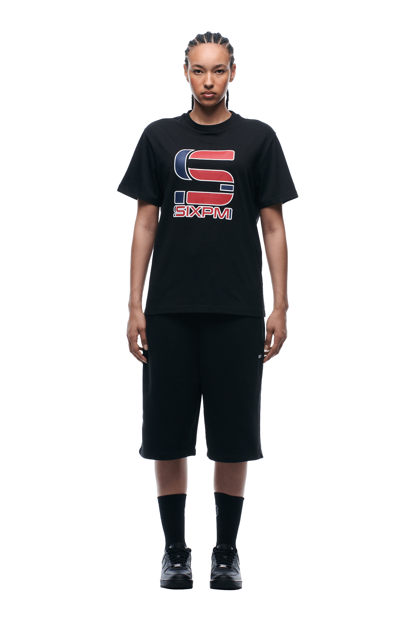 A person with cornrow braids stands facing forward, wearing the 6PM GRAN TURISMO TEE BLACK, paired with black knee-length shorts, black socks, and black sneakers on a plain background.