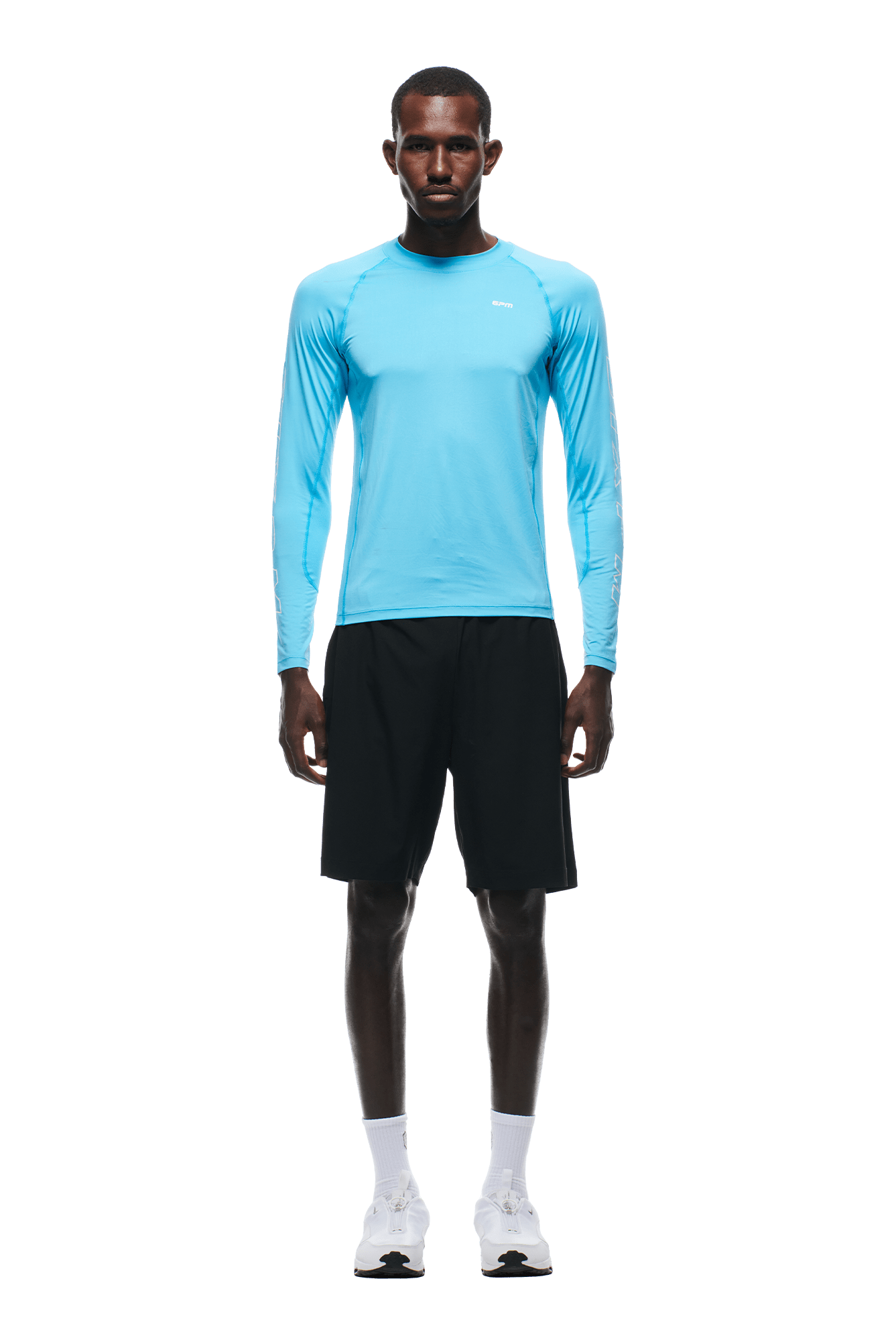A man stands facing forward against a plain background, wearing the 6PM SPORTS LONGSLEEVE AQUA BLUE shirt by 6PM, paired with black shorts, white socks, and white sneakers.
