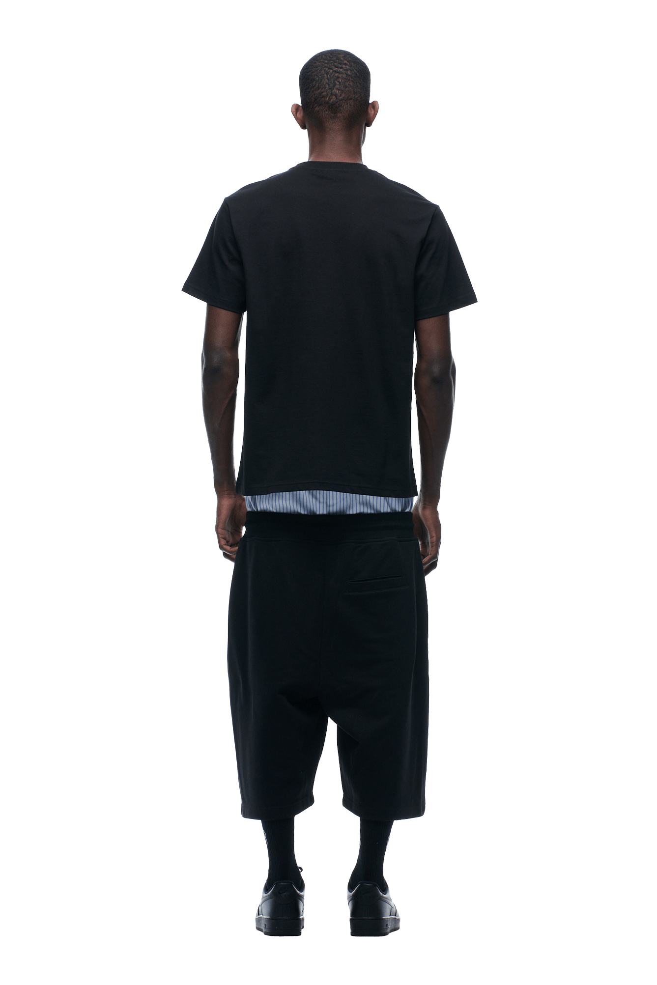 A person stands facing away in the 6PM GRAN TURISMO TEE BLACK, loose black shorts, black socks, and black shoes. A striped shirt hem peeks from under the tee. The background is transparent.