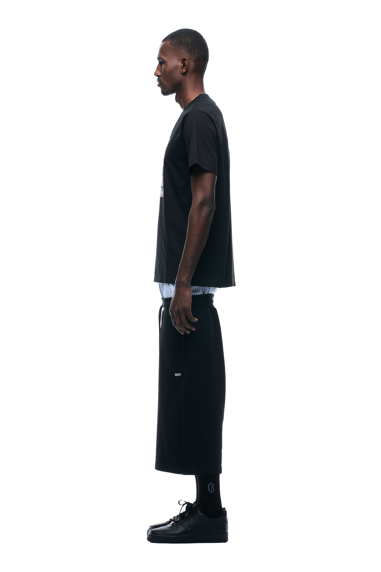 A man stands in profile facing left, wearing the 6PM GRAN TURISMO TEE BLACK with a black skirt, socks, and sneakers, set against a plain white background.