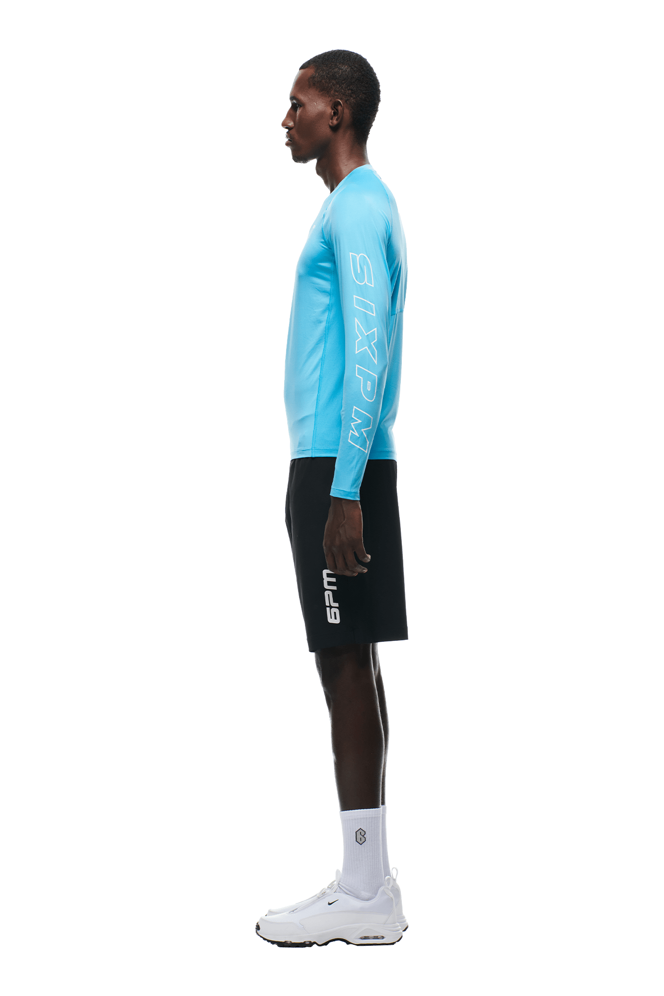 A person stands in profile against a plain background, wearing the 6PM SPORTS LONGSLEEVE AQUA BLUE shirt with SIXPM on the sleeve, black shorts with 6PM branding, plus white socks and sneakers.