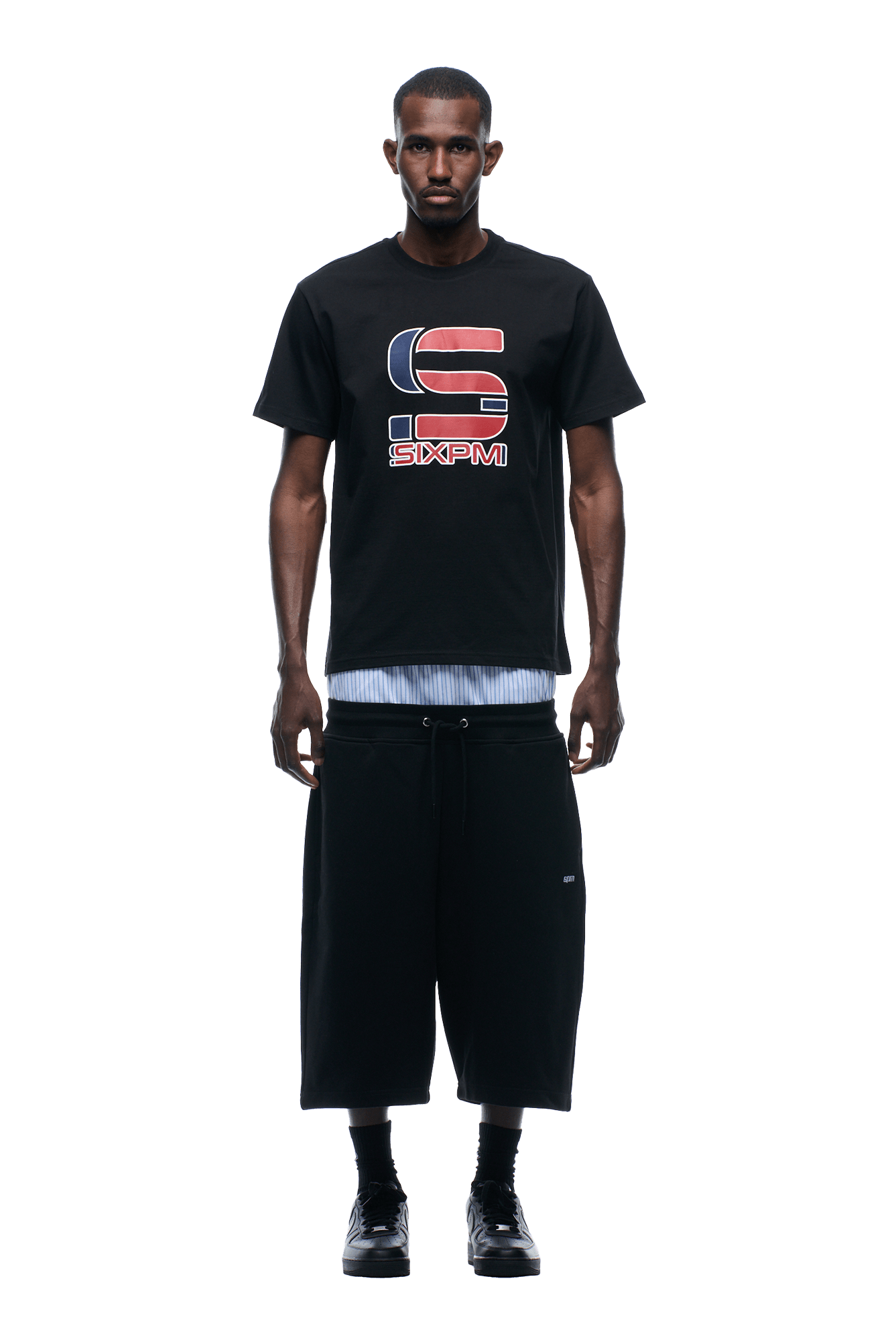 A man faces forward against a plain background, wearing the 6PM GRAN TURISMO TEE BLACK over a striped shirt, paired with loose black knee-length shorts and black shoes.